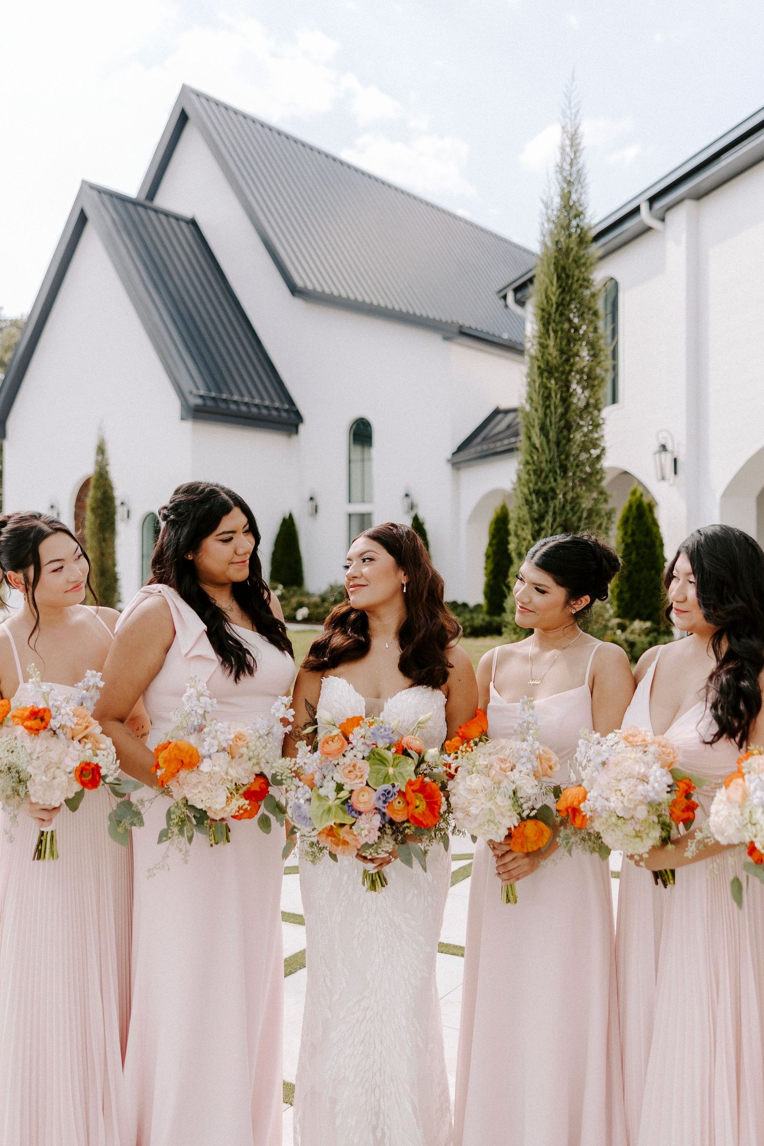Bridal party wearing blush pink dresses holds vibrant orange and white floral bouquets outside The Grand at Willow Springs.