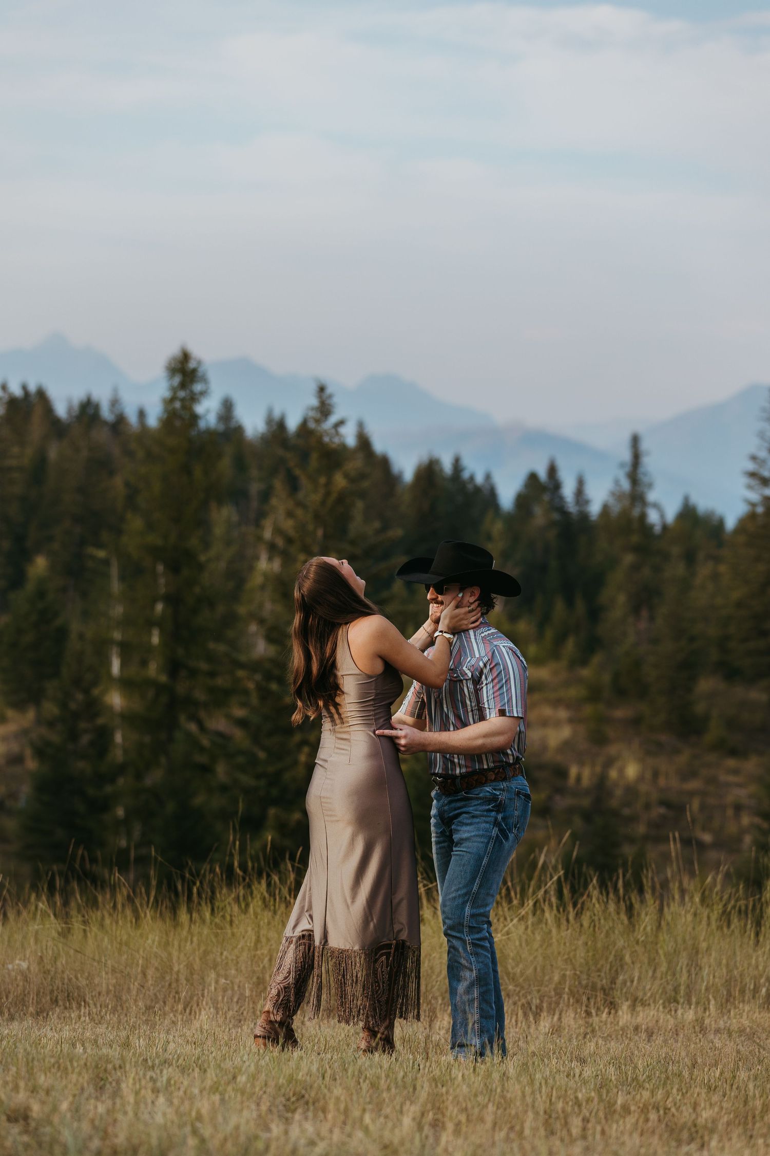 A couple in western attire dancing together in a meadow with mountains and forest in the background at dusk.