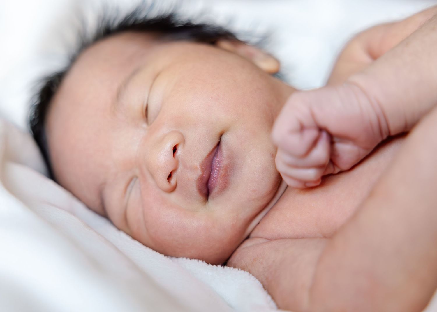 A sleeping newborn baby with closed eyes and peaceful expression rests against soft white bedding.
