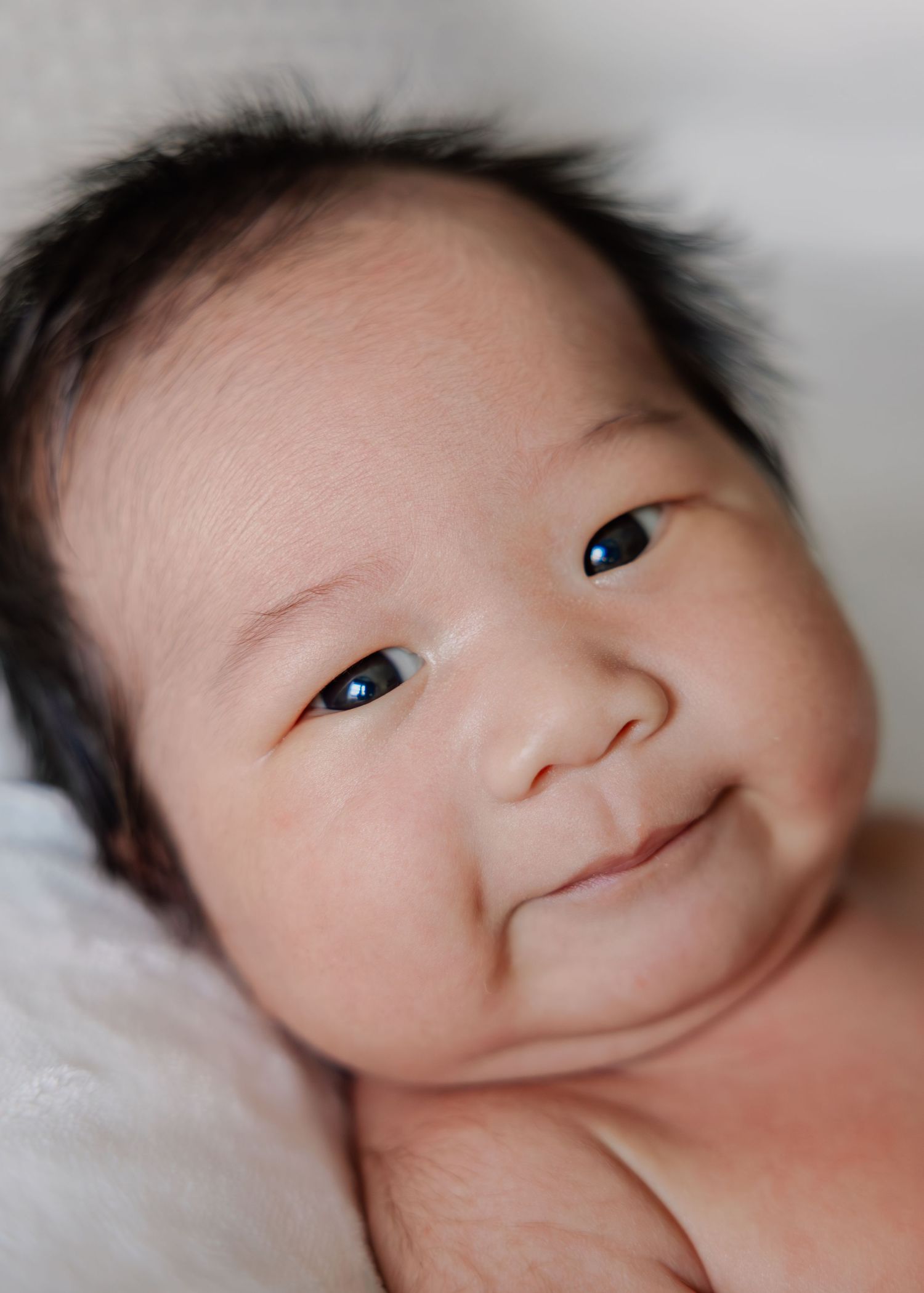 A newborn baby with dark hair rests on white bedding while making various adorable facial expressions.