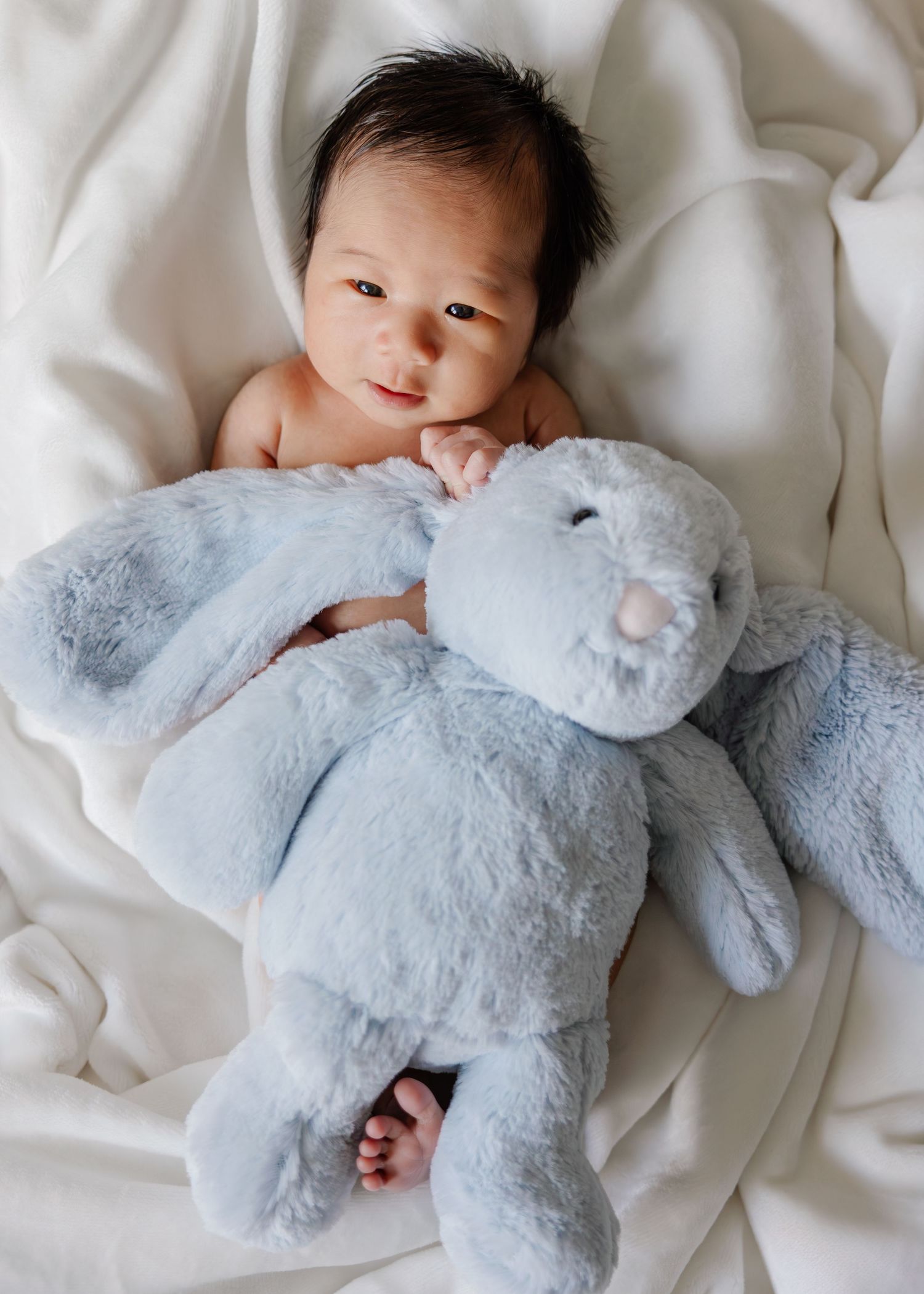 A baby lies on white bedding while cuddling with a soft blue plush bunny toy.