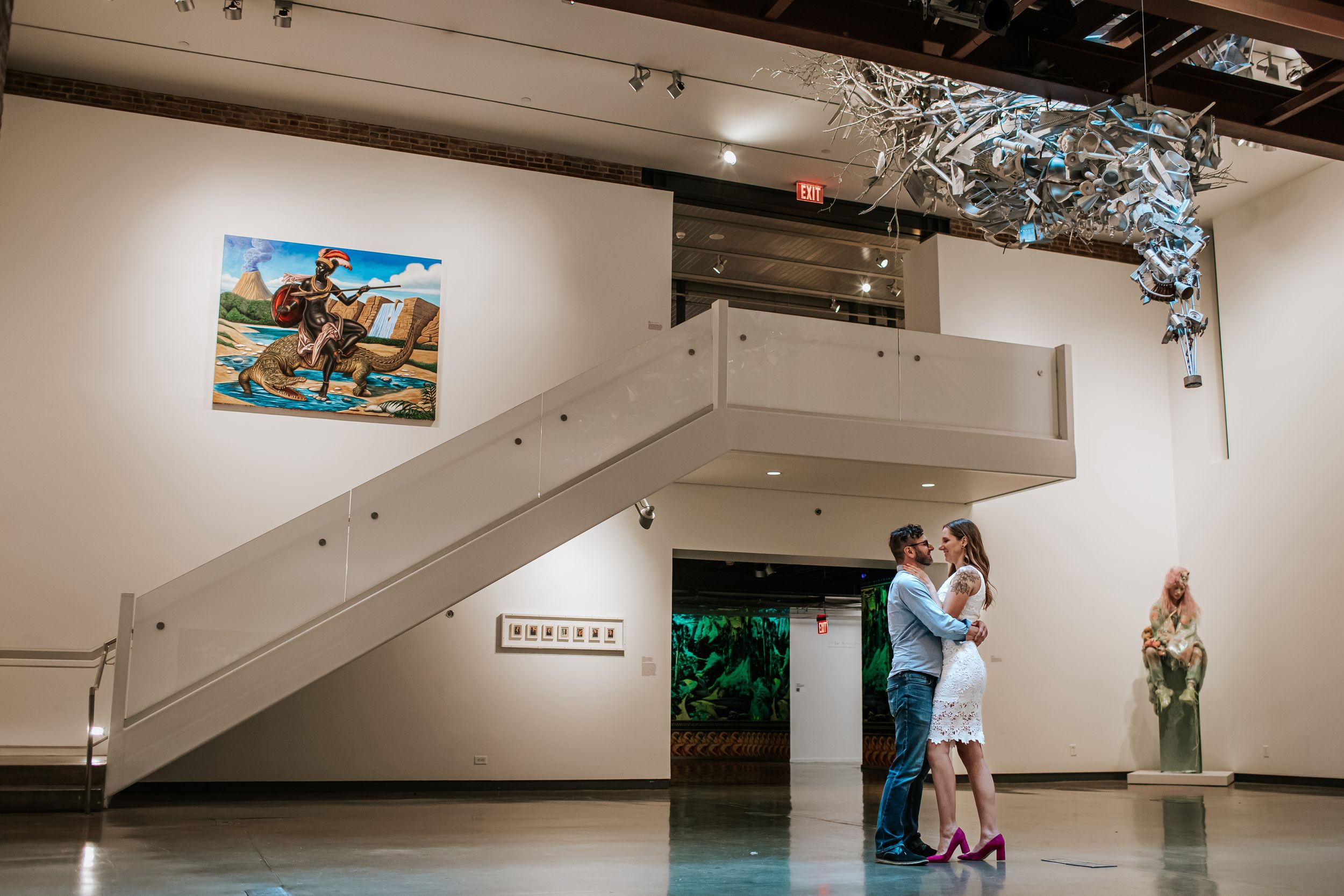 A modern art gallery space with white walls displays framed artwork and features suspended decorative elements from the ceiling.