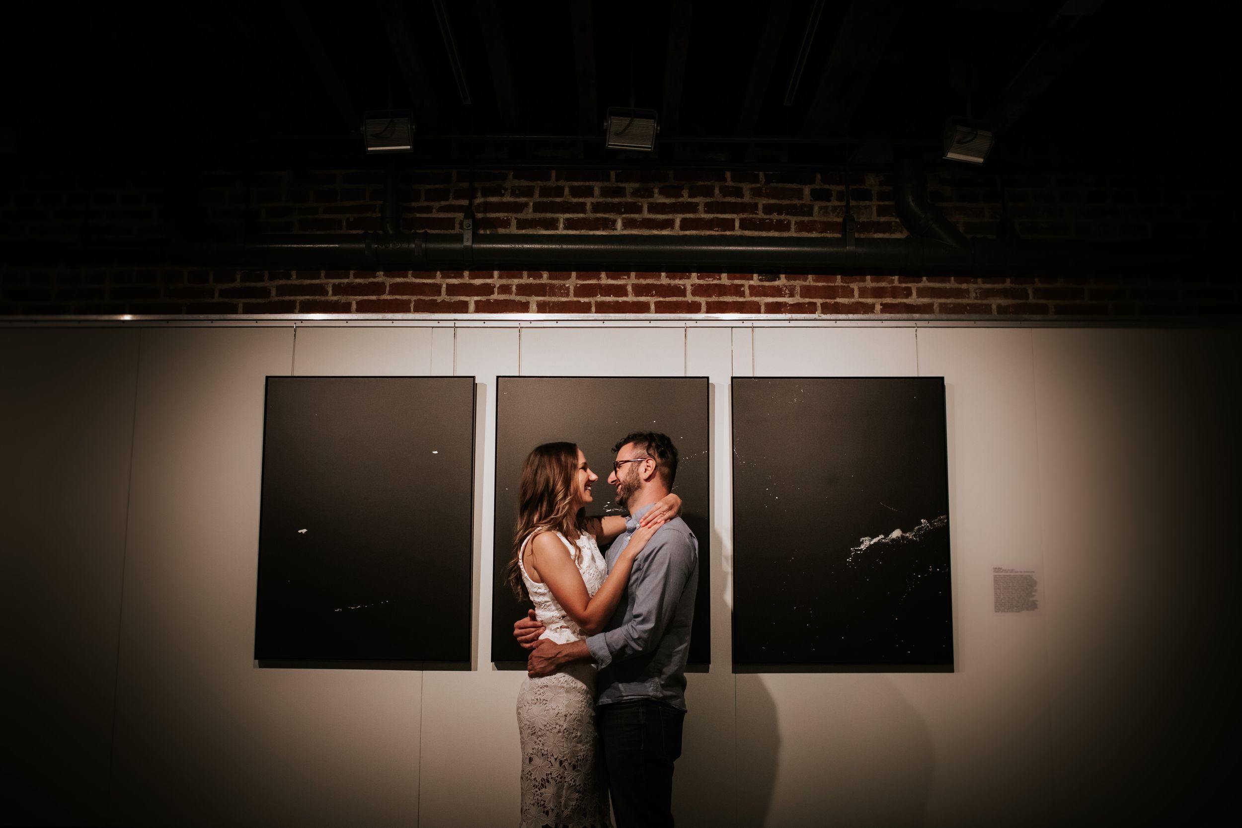 A romantic couple embraces in front of three dark panels in a dimly lit art gallery.