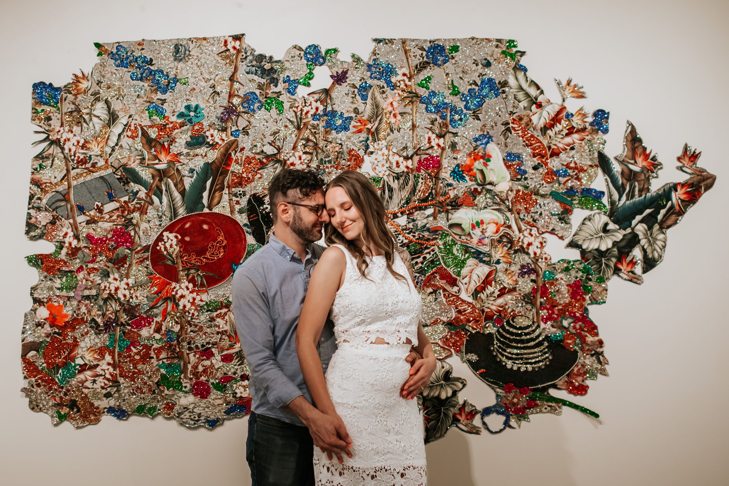A couple poses together in front of a vibrant wall art installation made of colorful butterflies and floral elements.