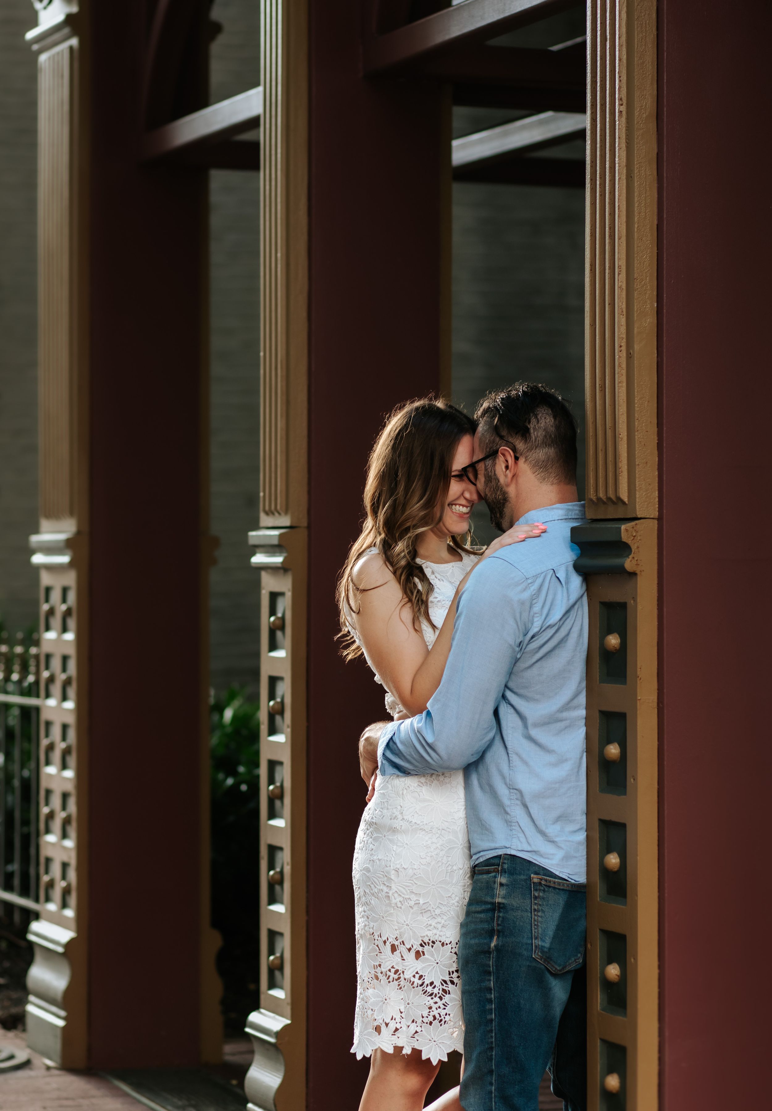 Couple embracing in a covered walkway with wooden columns and natural lighting.