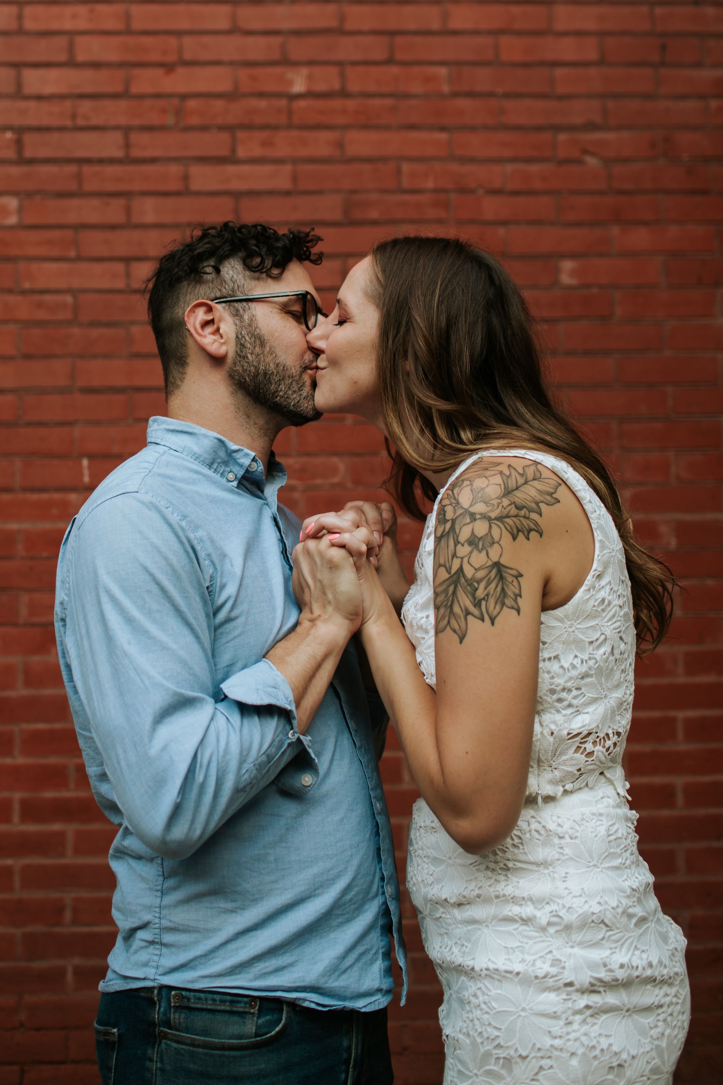 A couple embraces tenderly against a rustic red brick wall backdrop.