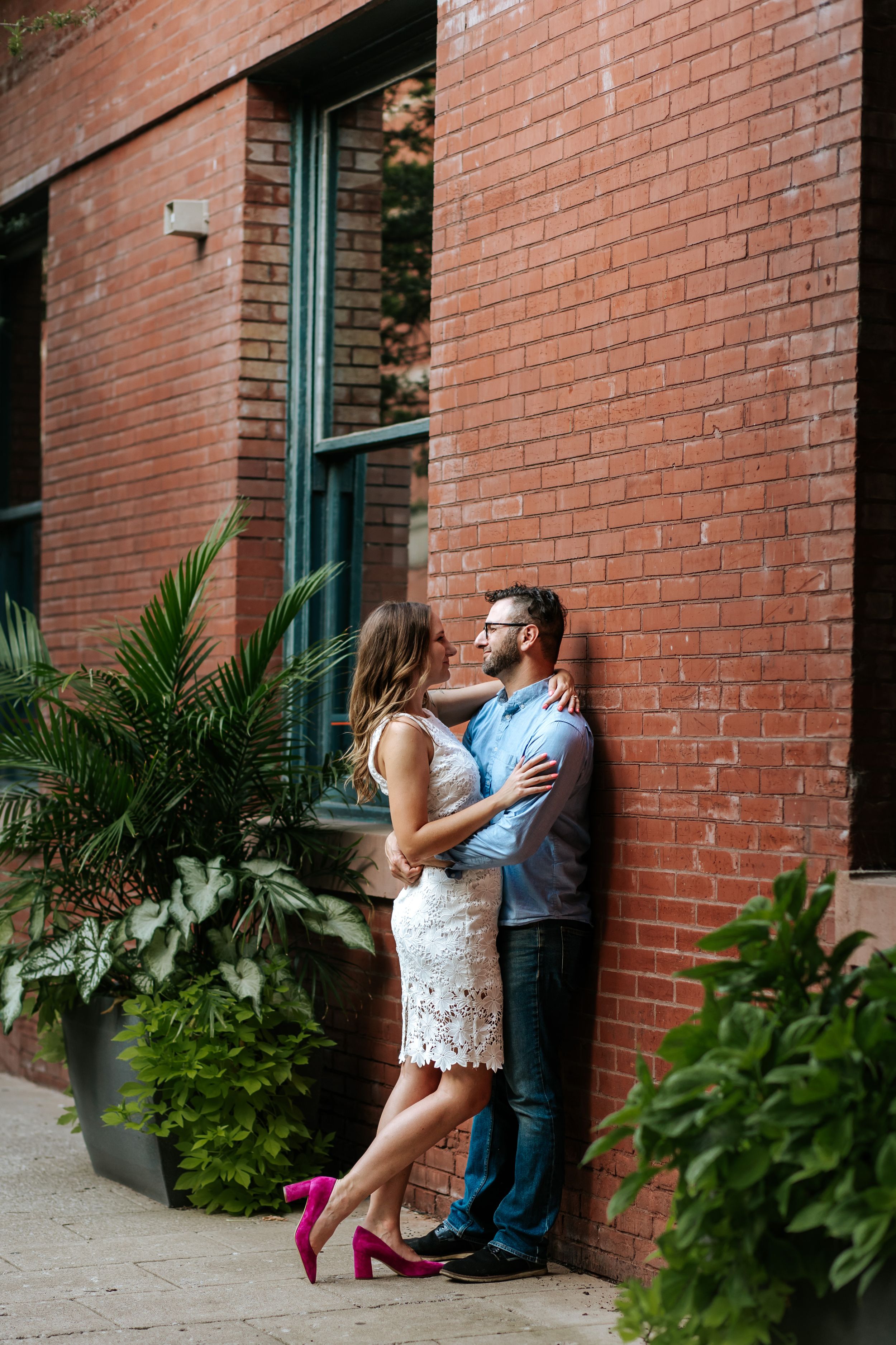 Romantic moment against red brick wall with lush green plants and bright magenta shoes.