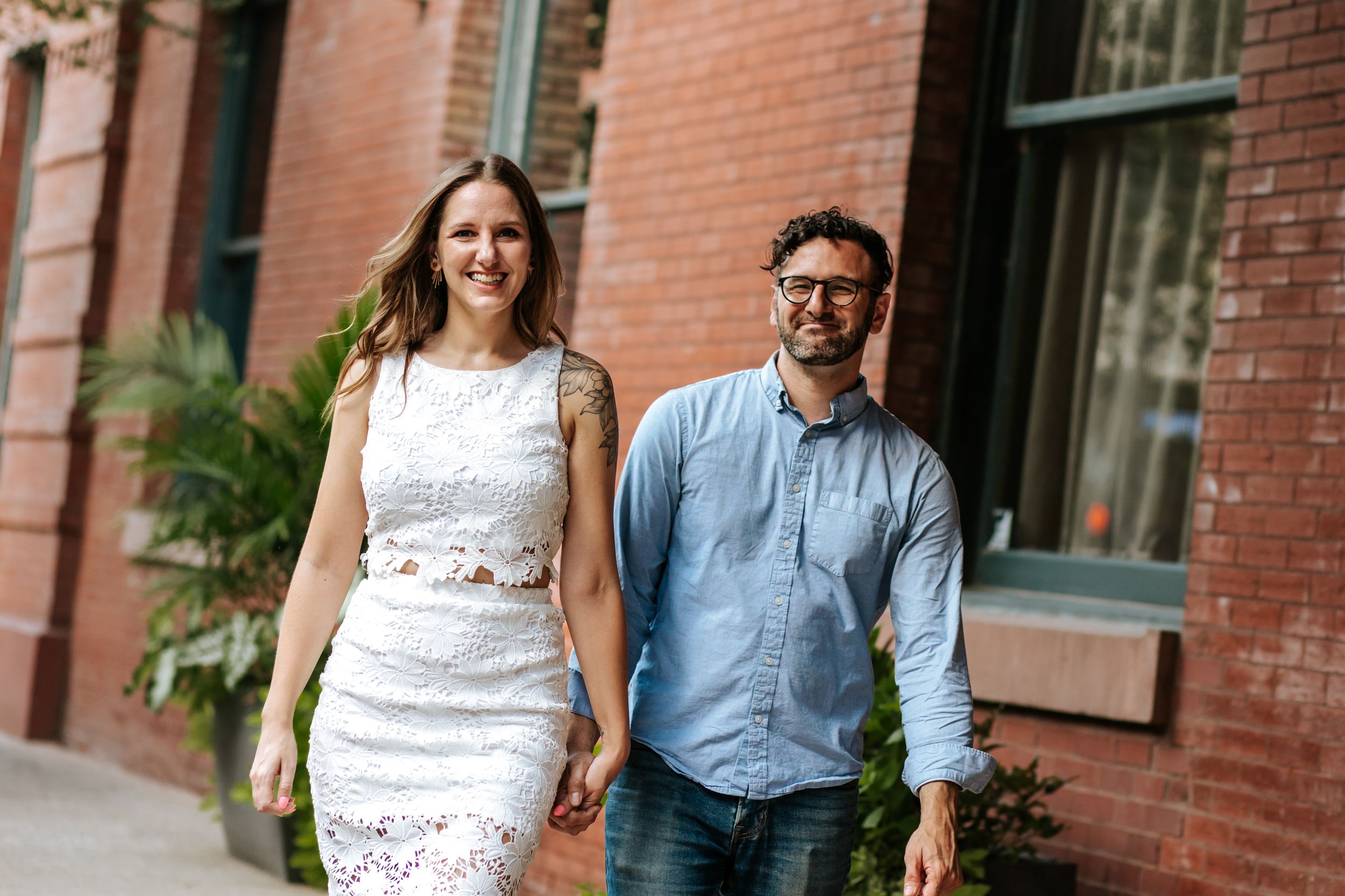 A couple walks together smiling on a sidewalk by a brick building with ferns in the background.