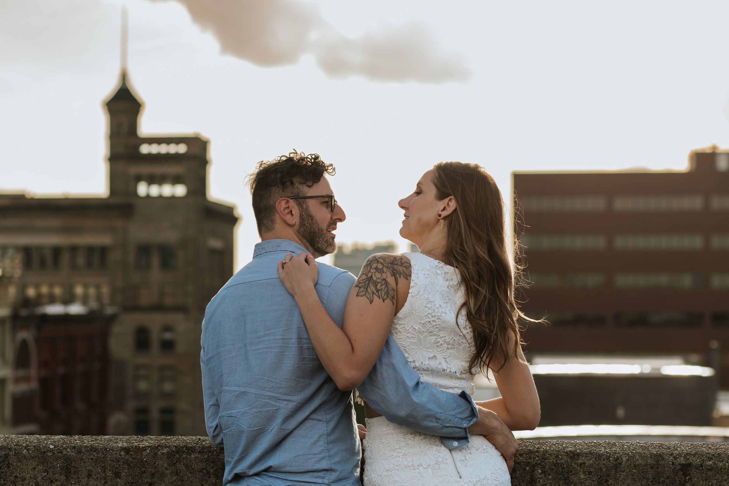Couple sits on stone wall overlooking city skyline at sunset while sharing intimate moment.