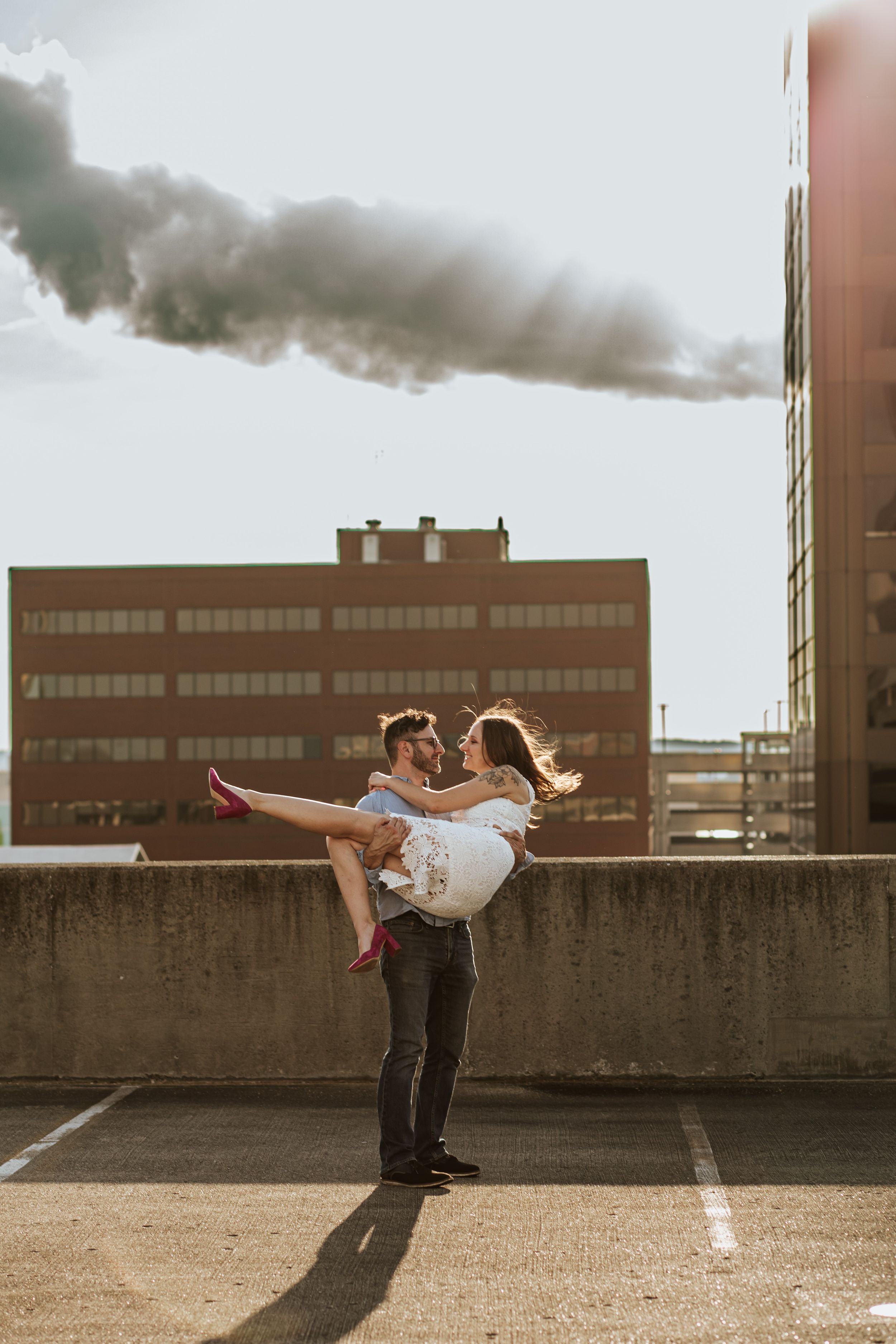 A couple shares a romantic moment on a rooftop parking garage at sunset with dark smoke billowing in the background.