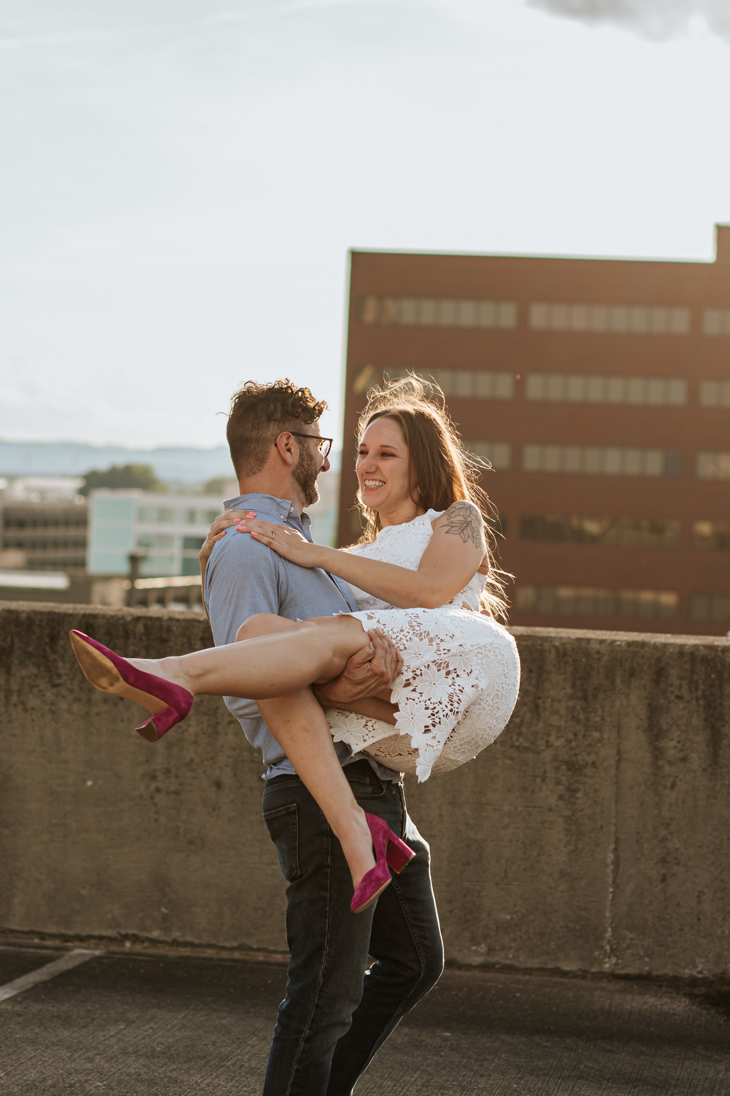 Couple dancing together on a rooftop at sunset, with the woman wearing a white lace dress and pink heels.