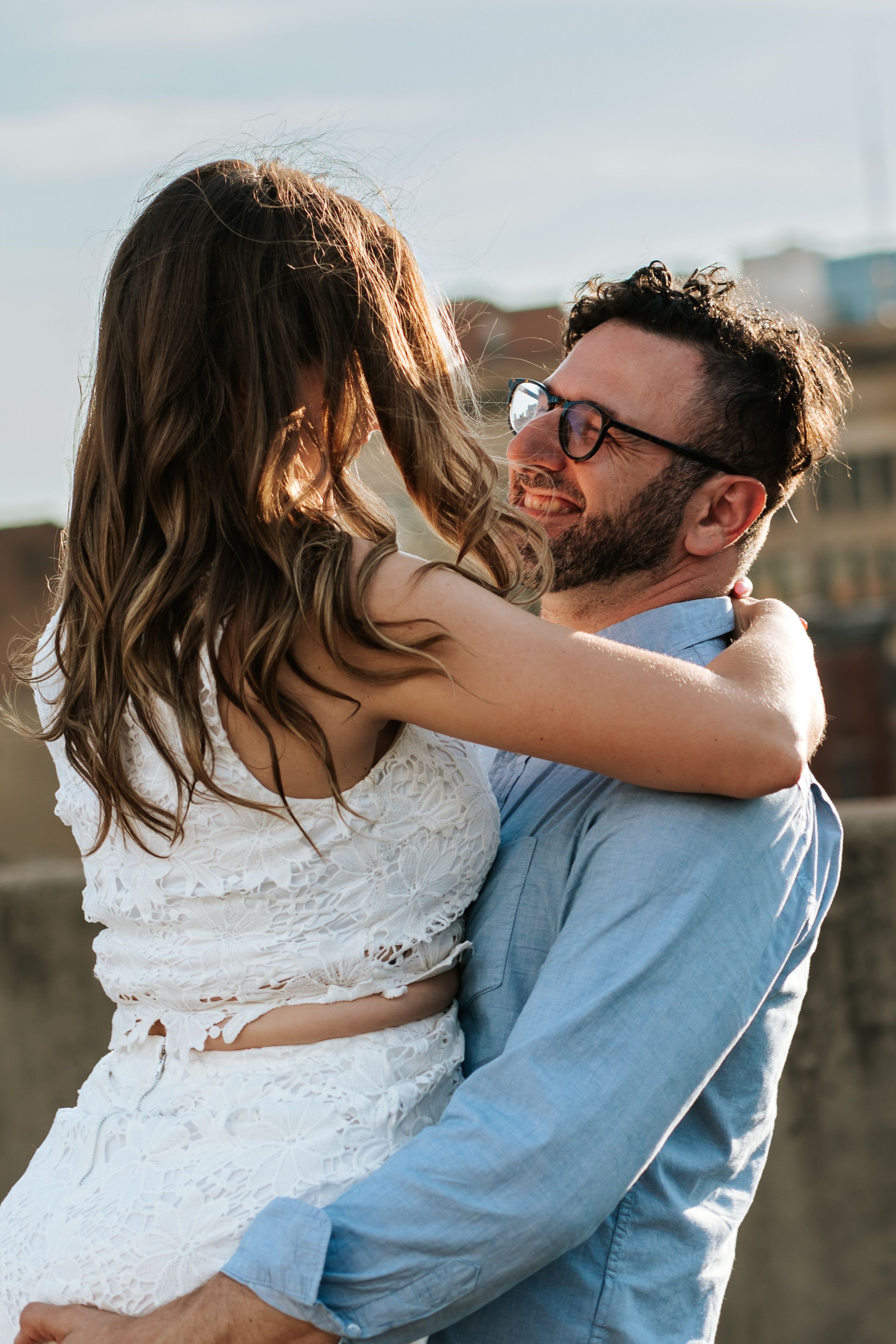 A couple embraces outdoors in wedding attire, with the bride in white lace and groom wearing a light blue shirt and glasses.
