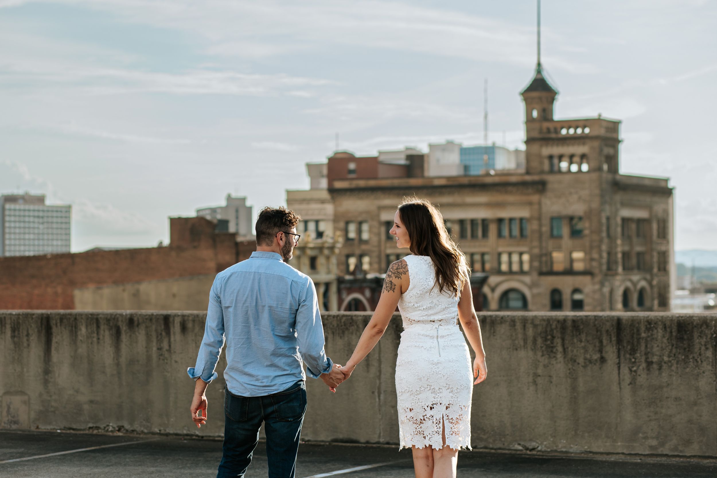 Couple walking hand in hand along city rooftop with historic architecture in background.