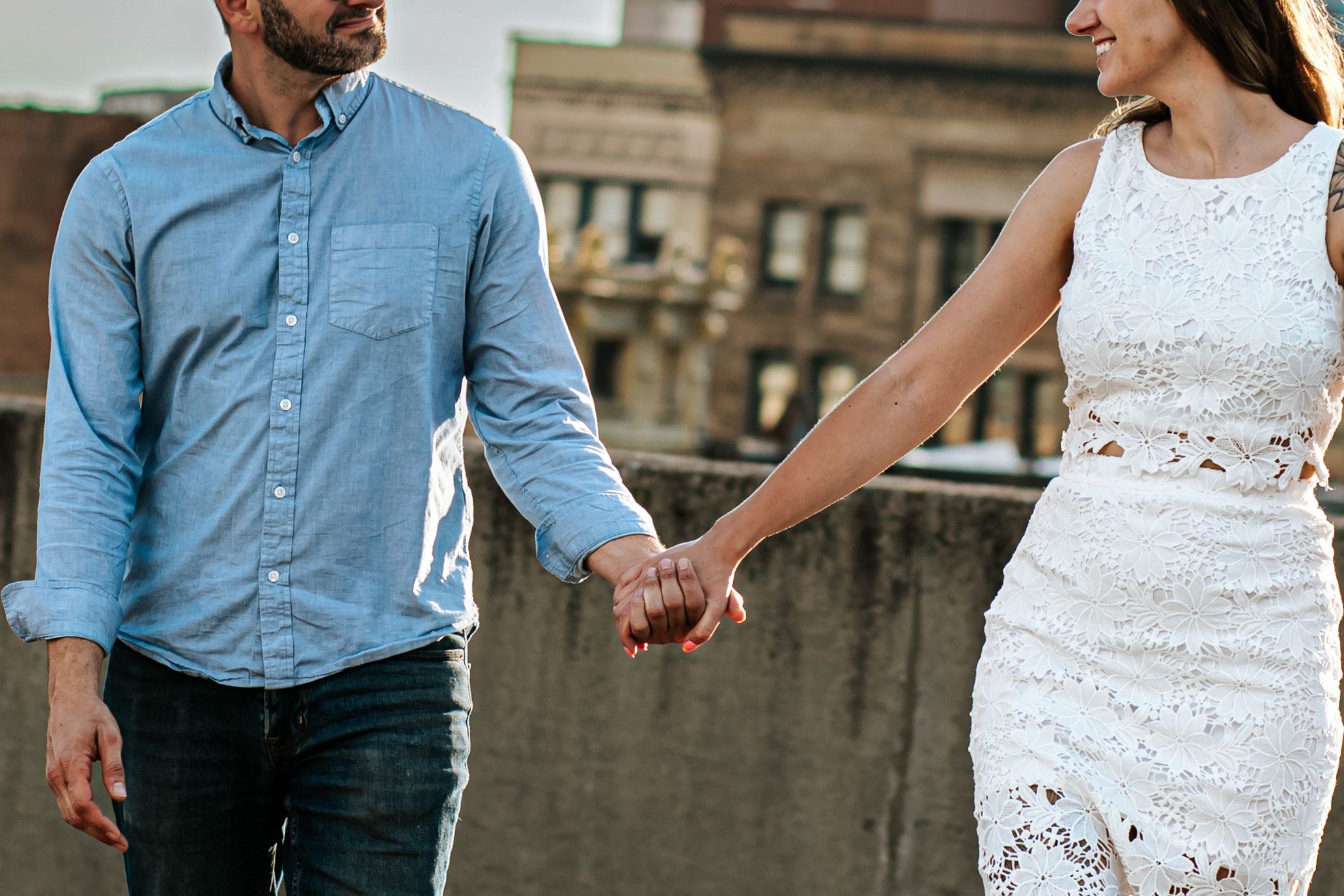 Couple in blue shirt and white lace dress holding hands against urban rooftop setting.