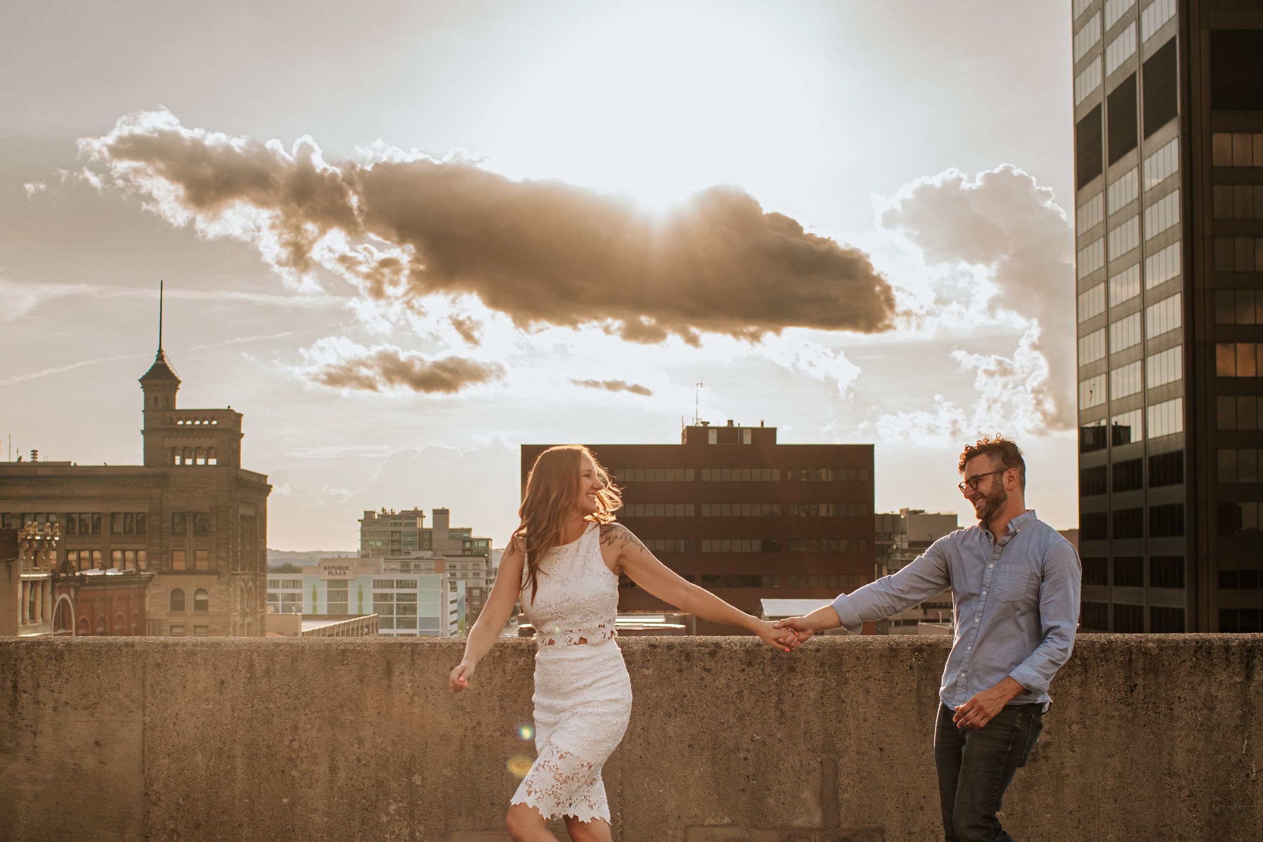 Two people share a joyful moment on a rooftop with golden sunlight and city buildings in the background.