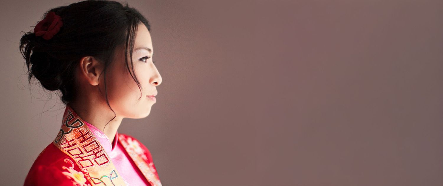 Asian bride in profile wearing traditional red Chinese qipao dress against neutral background.