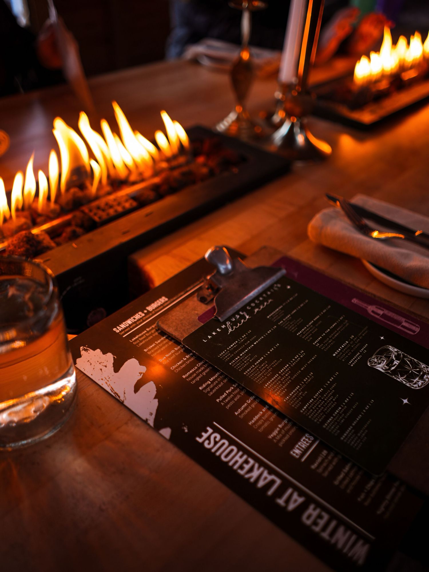 A restaurant menu sits on a dark wooden table illuminated by warm candlelight with a cocktail glass nearby.