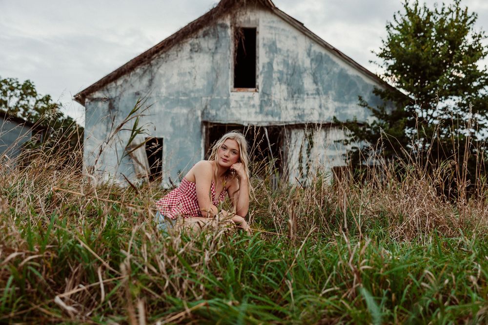 Someone in a pink dress sits in tall grass in front of a weathered white farmhouse on an overcast day.