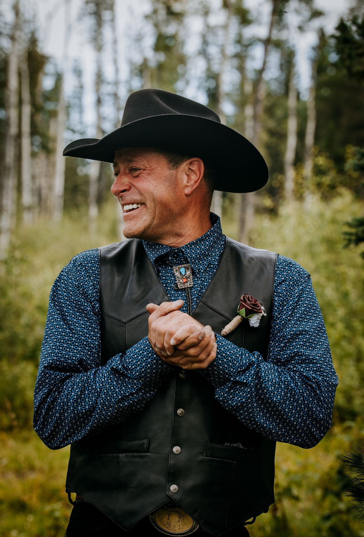 A person in western attire including black cowboy hat, polka dot shirt and leather vest smiles while standing outdoors.