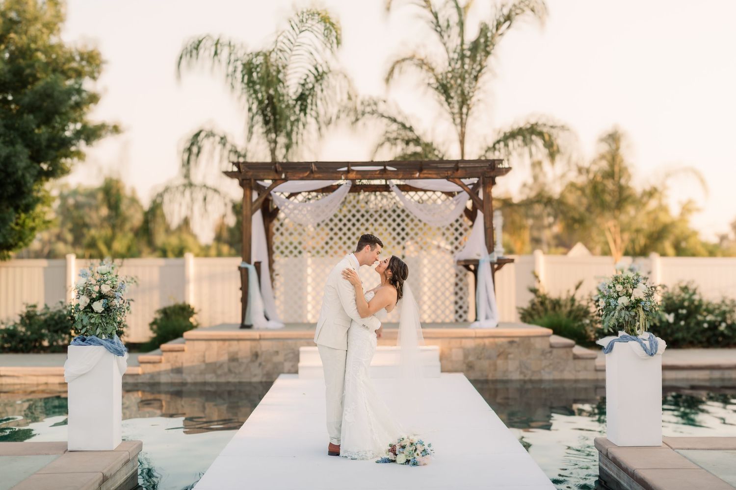 A romantic wedding ceremony setup with white chairs and palm trees against a golden sunset backdrop.