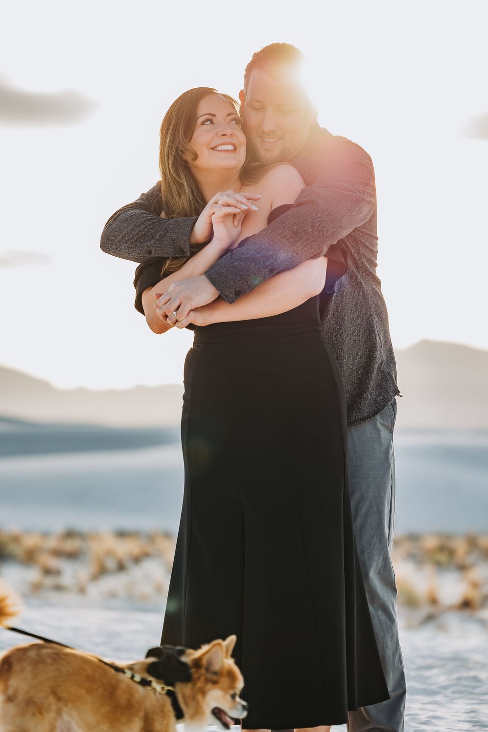 A couple embraces on a rocky beach at sunset with their dog watching nearby.