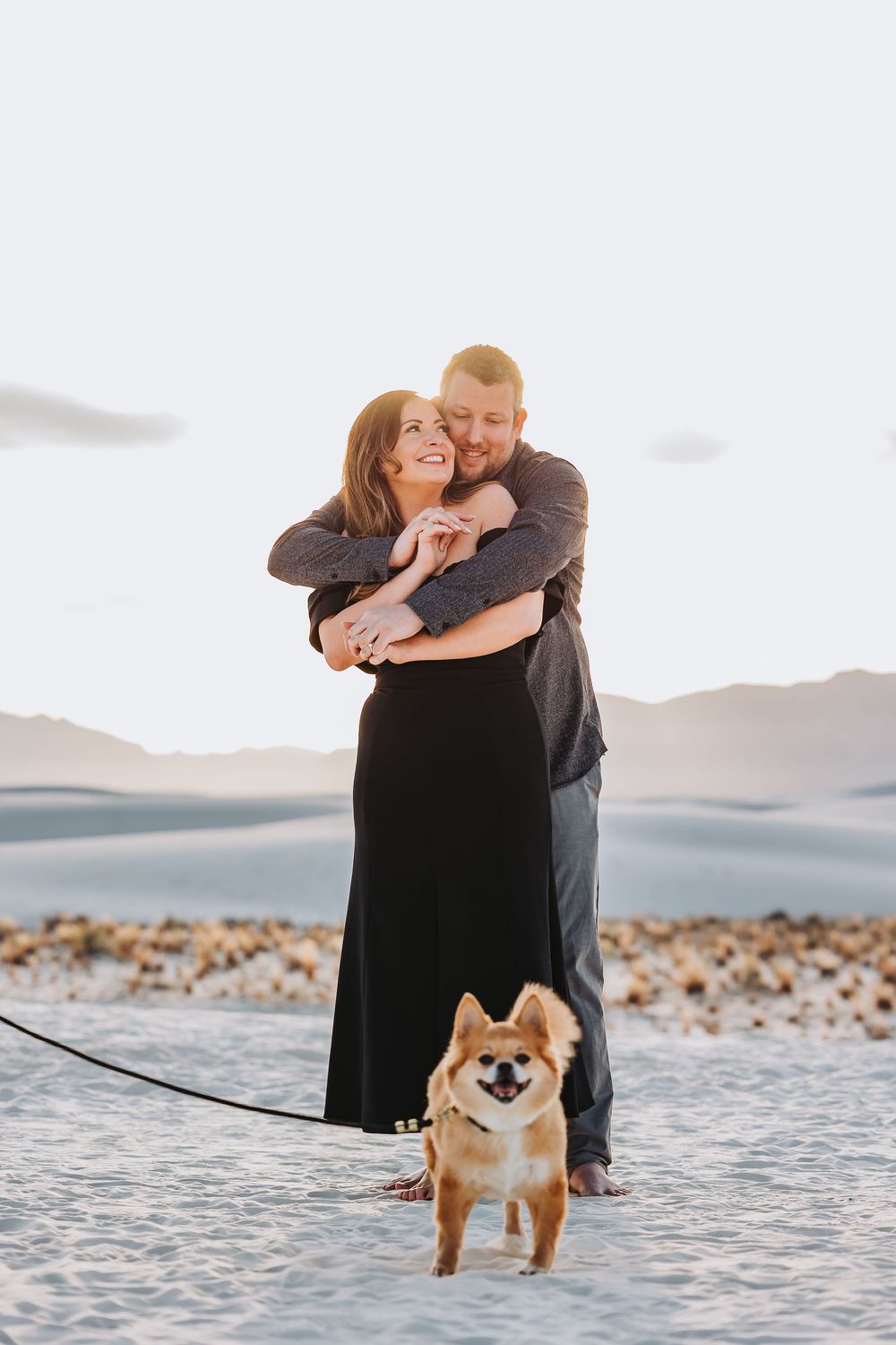 A couple embraces on a white sandy beach with their Shiba Inu dog during sunset.