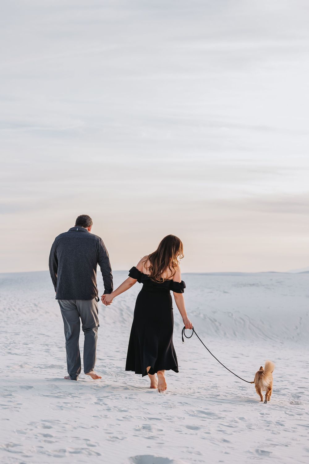 Couple walking their small dog together on pristine white sand dunes at sunset.