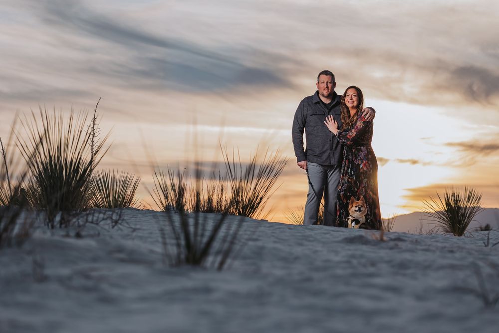 A couple embraces on a sandy beach dune during golden hour sunset as the sky glows warmly behind them.