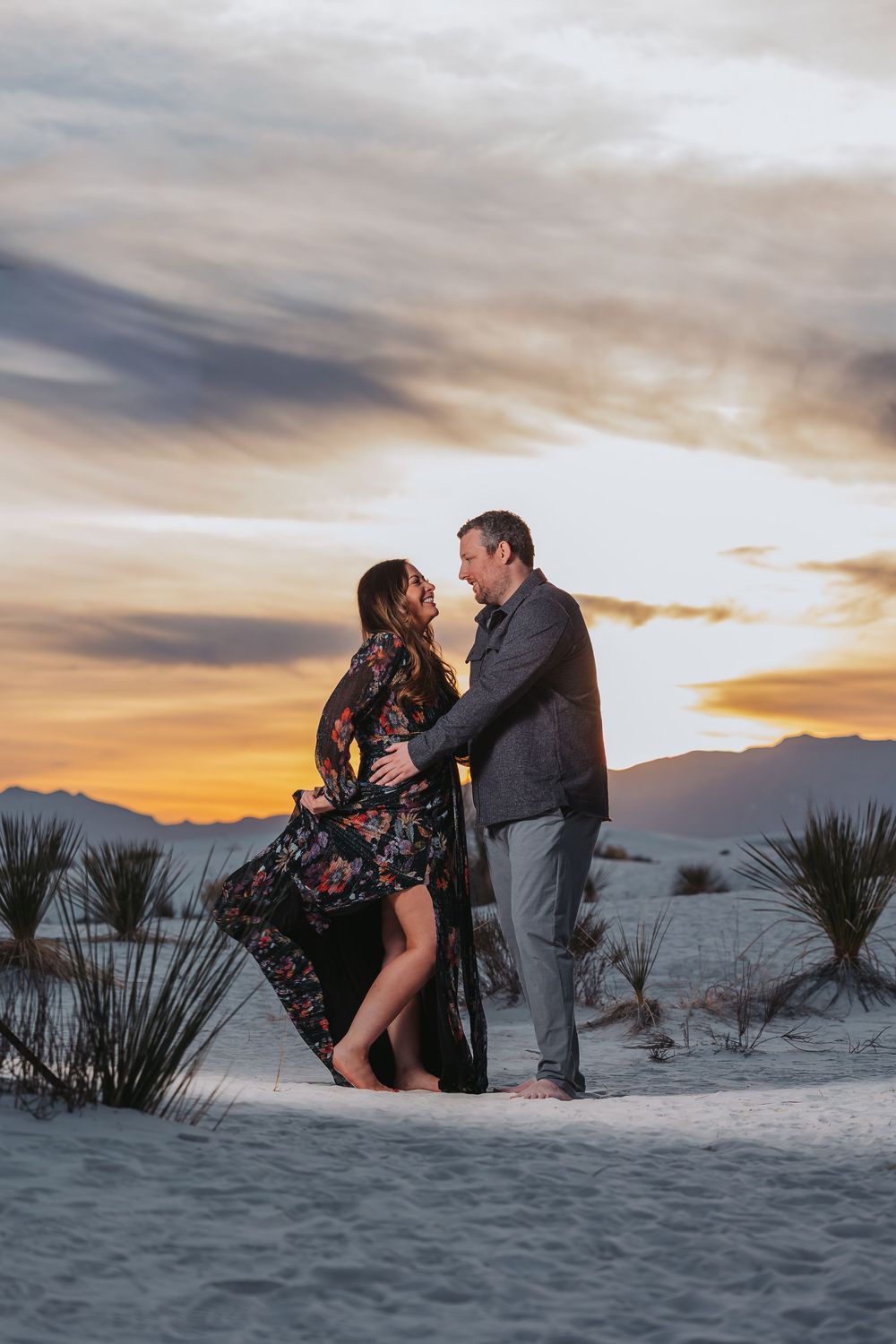 A romantic couple embraces on white sand dunes during a dramatic desert sunset with mountains in the background.