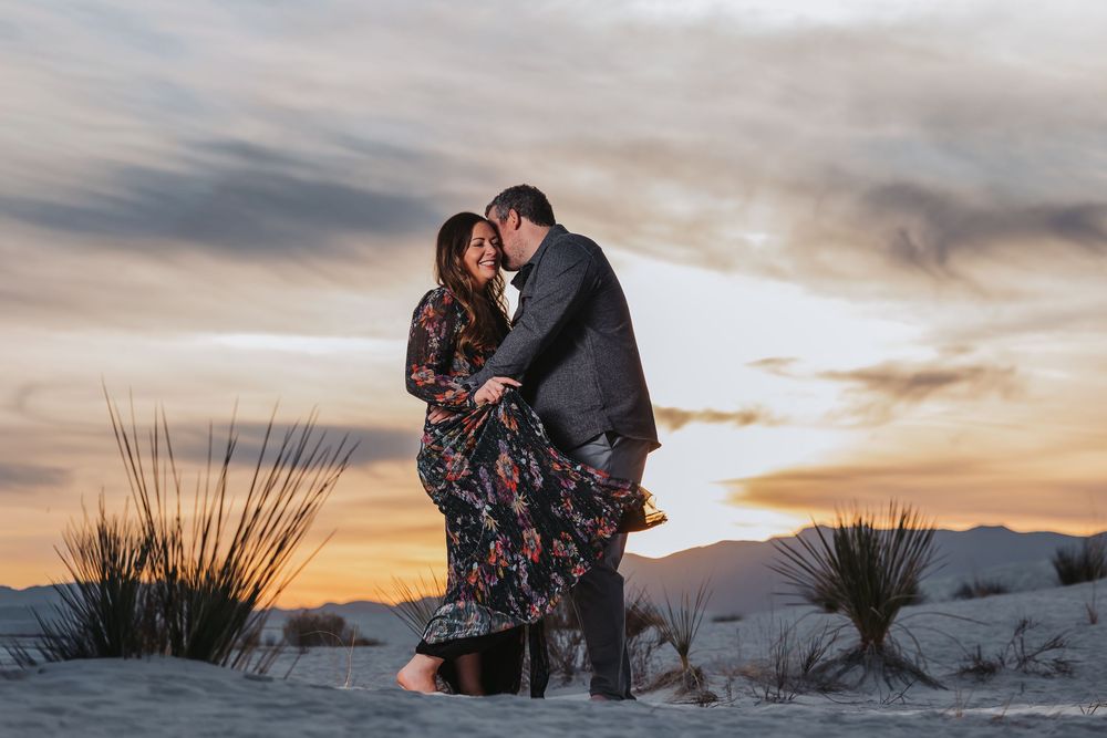 Two people embrace in a romantic moment at White Sands National Park during a dramatic desert sunset.