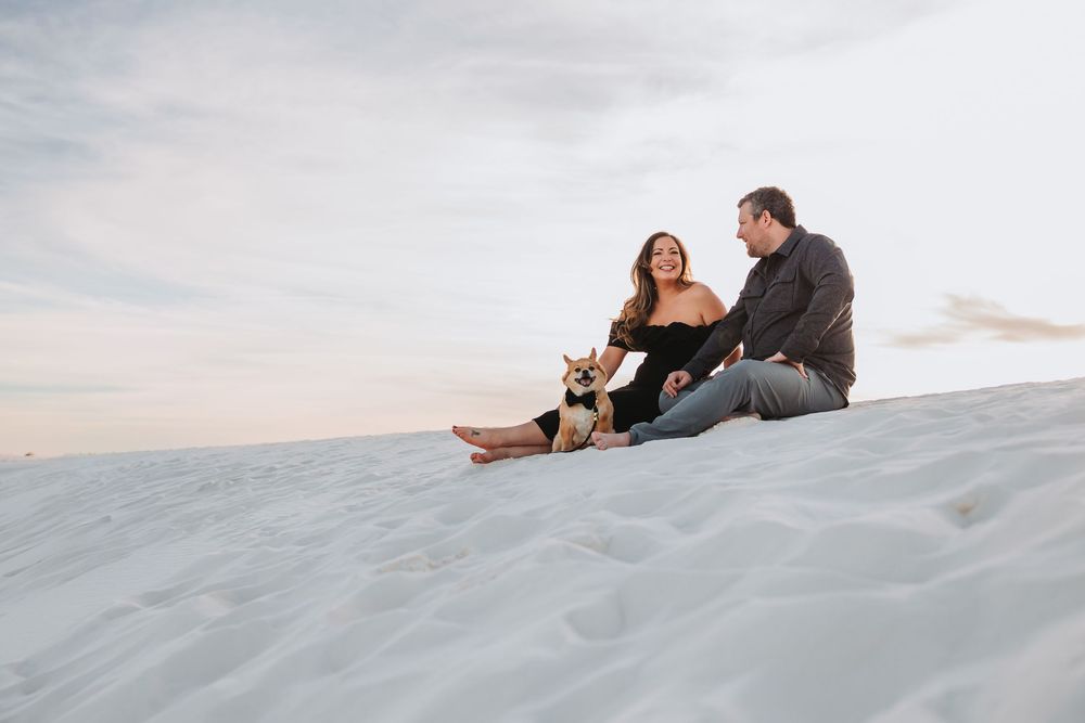 Couple sits together on white sand dunes at sunset creating silhouettes against the sky.