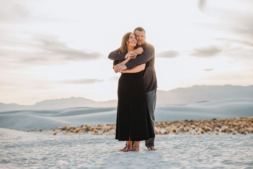 A couple embraces while standing in shallow water with mountains and cloudy skies in the background.