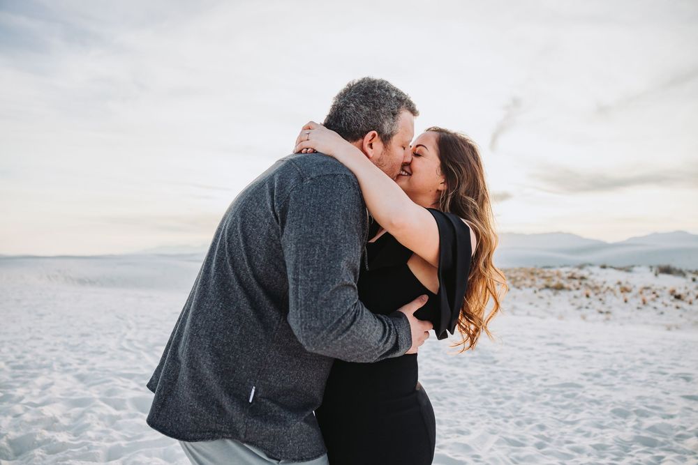 A romantic couple embraces and kisses on a snowy winter beach with mountains in the background.