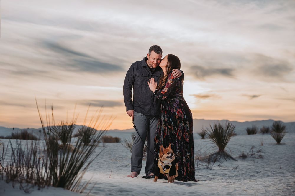 A couple shares a romantic moment at sunset in White Sands National Park with their corgi dog beside them.