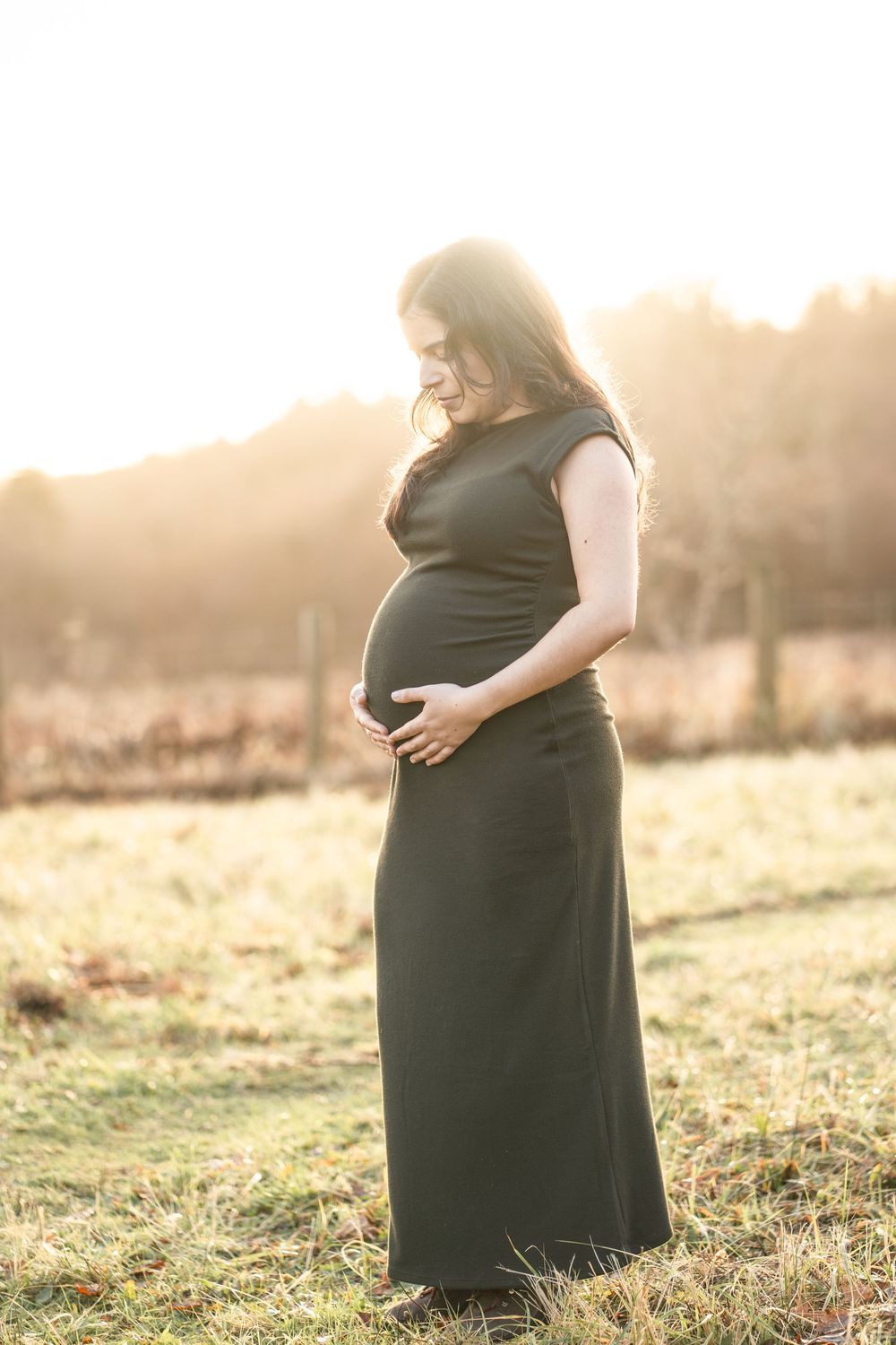Silhouette of pregnant woman in black dress standing in golden sunlit field during sunset.