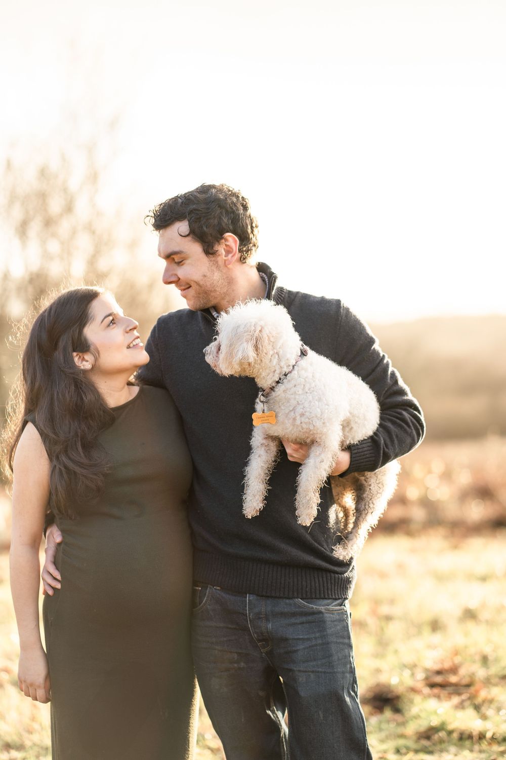 A couple holds their small white dog while sharing tender moments in a sunlit outdoor setting during golden hour.