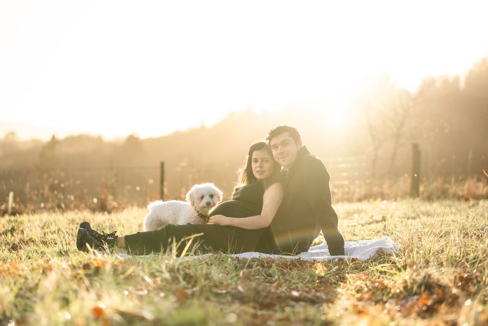 A couple relaxes on a grassy hill during golden hour sunset with their dog beside them.