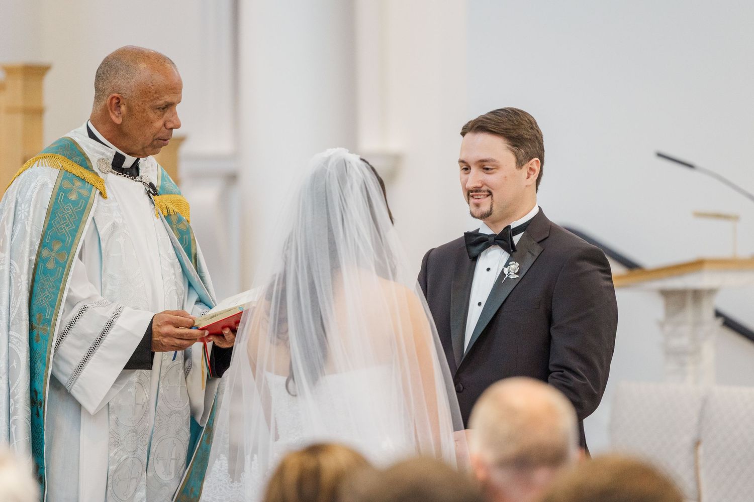 A couple exchanges vows during a wedding ceremony as a priest officiates in a bright, elegant church setting.
