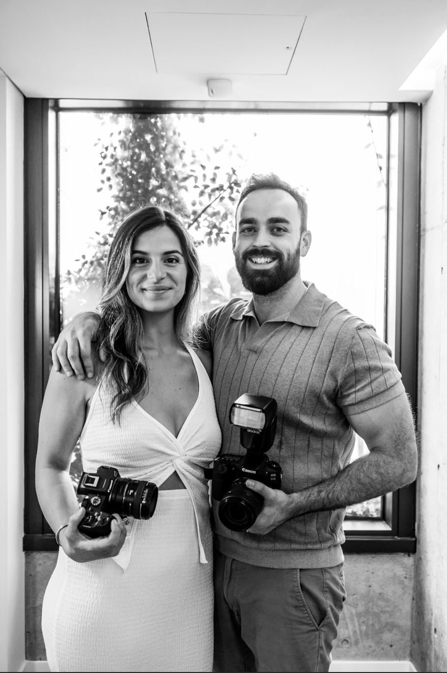 Black and white photo of a smiling couple holding professional cameras, posing together indoors near a window.