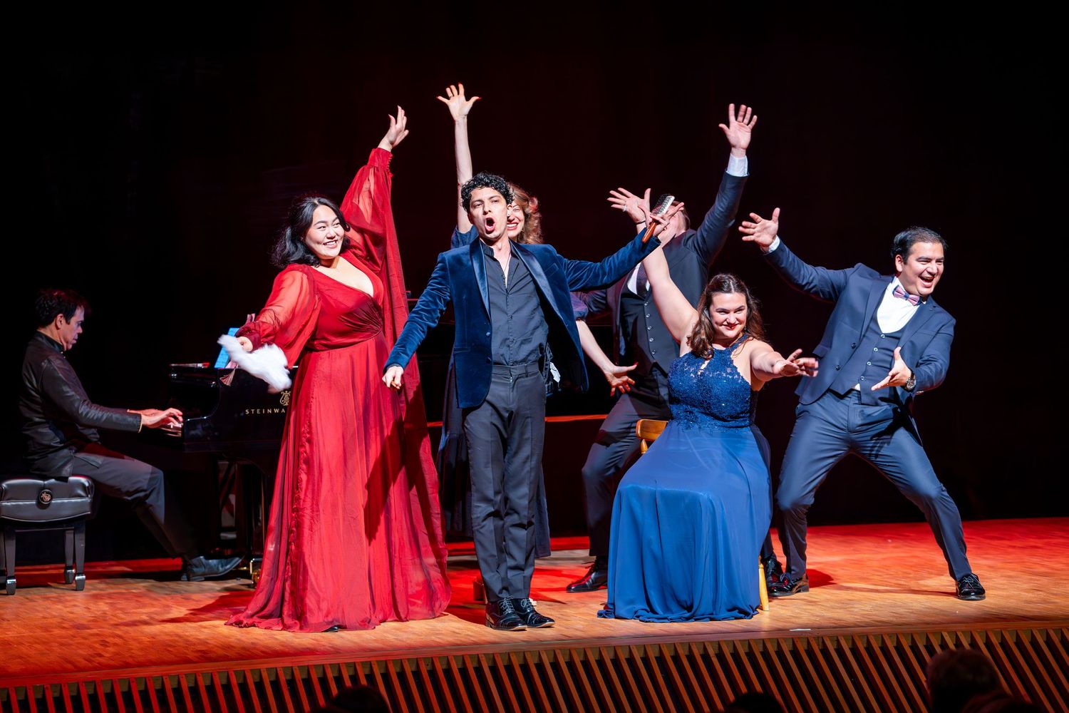 Performers in red and blue dresses dance energetically on a theater stage during a dramatic scene.