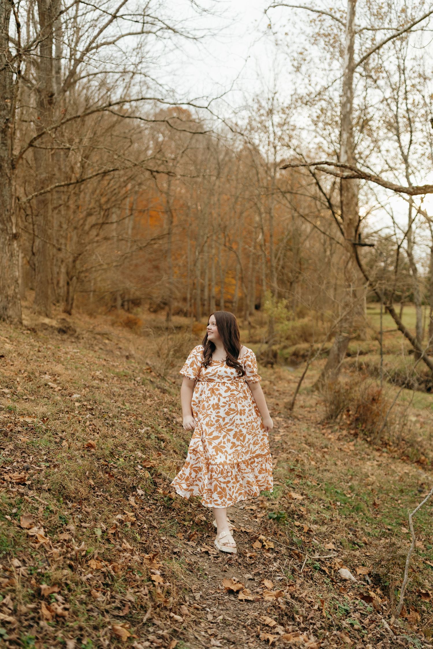 A figure in a floral dress walks through a wooded autumn path covered in fallen leaves.