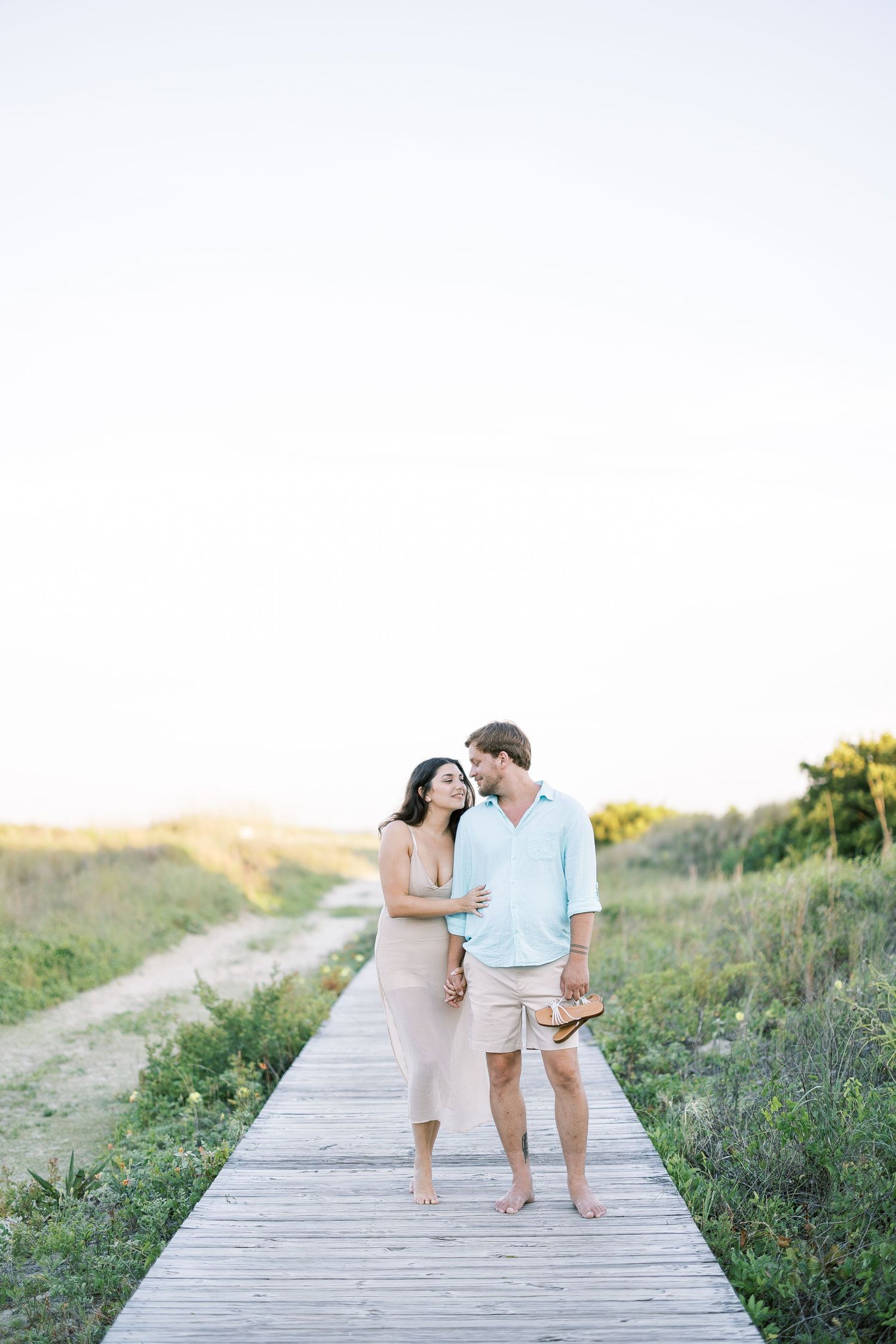 A couple in light summery outfits walks intimately along a wooden boardwalk through a seaside dune at golden hour.