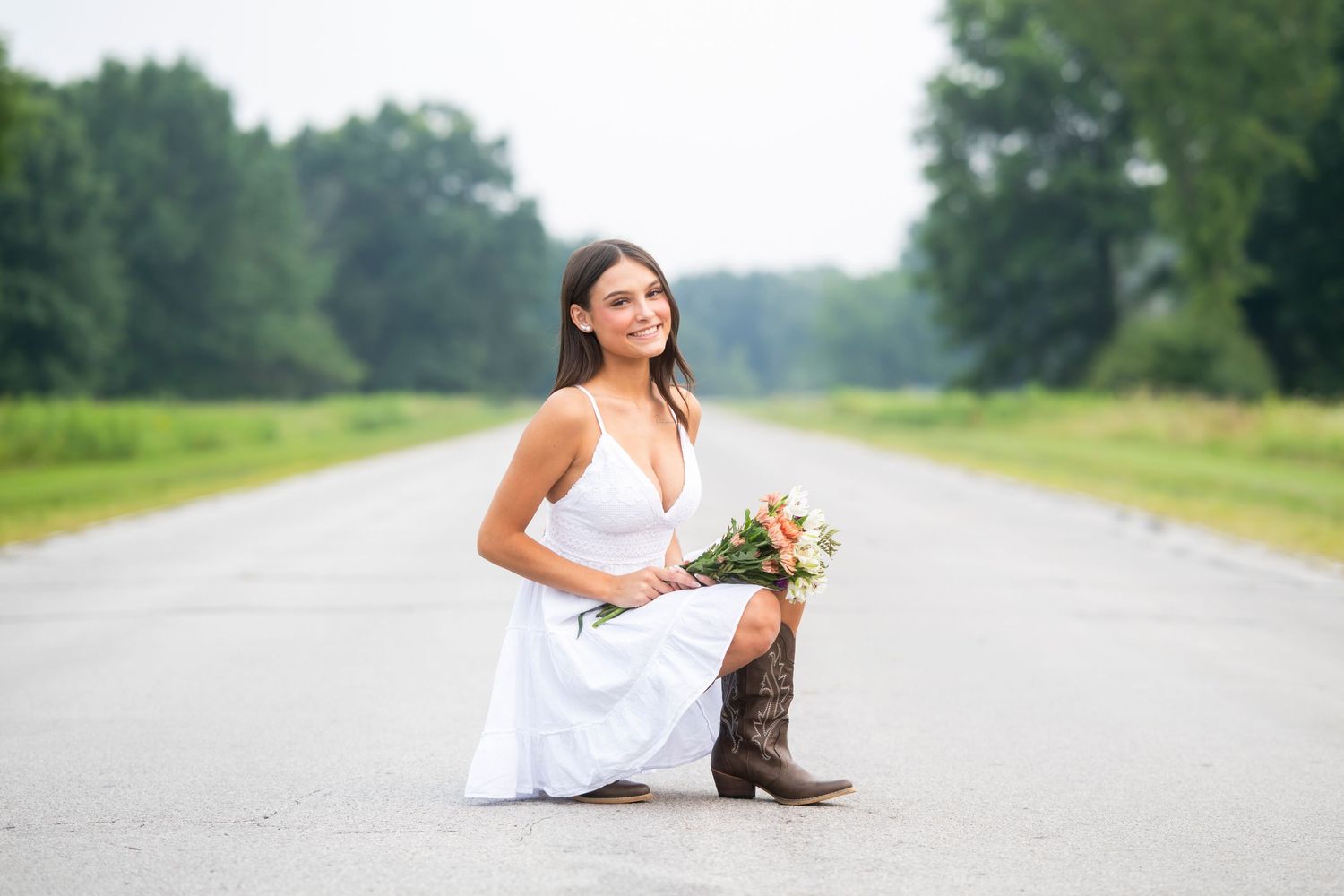 high school senior girl squatting down with a dress on and cowboy boots holding flowers
