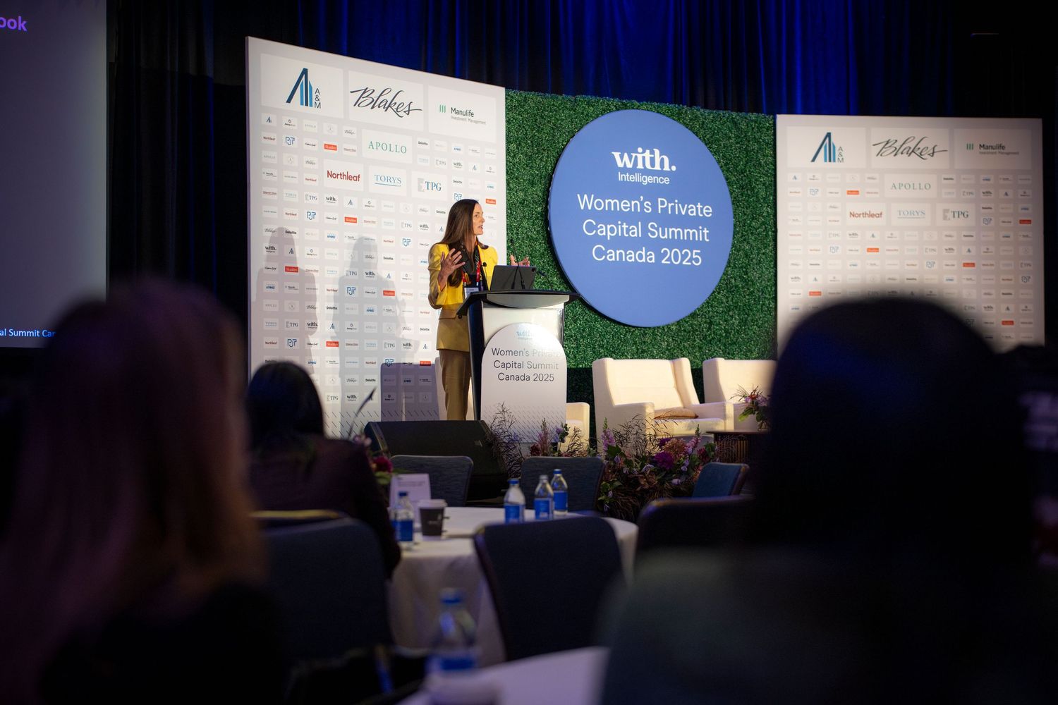 A speaker in a bright yellow blazer gestures while presenting at a business conference with branded backdrop panels.