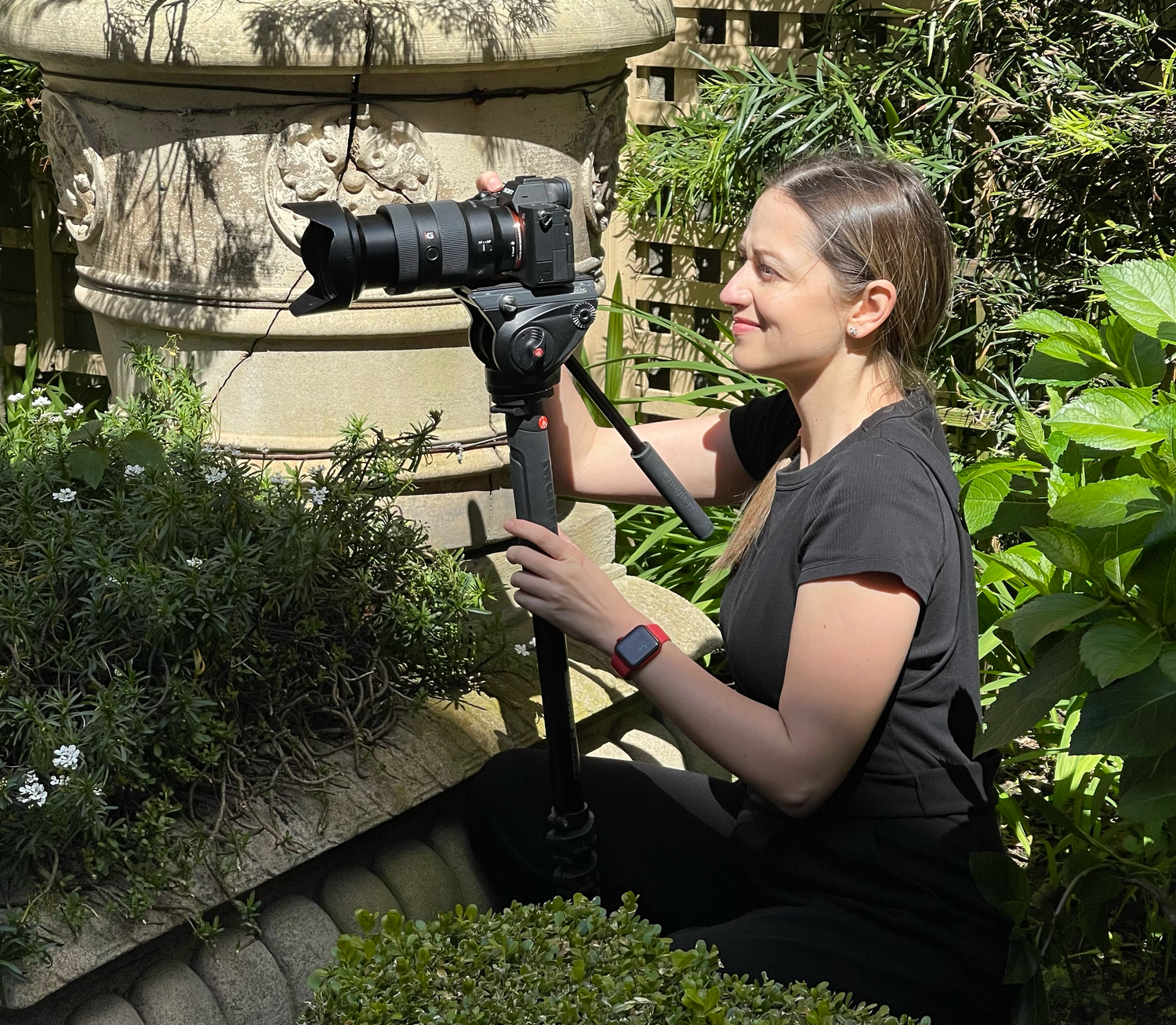 A photographer in black attire captures nature shots with a telephoto lens in a lush garden setting.
