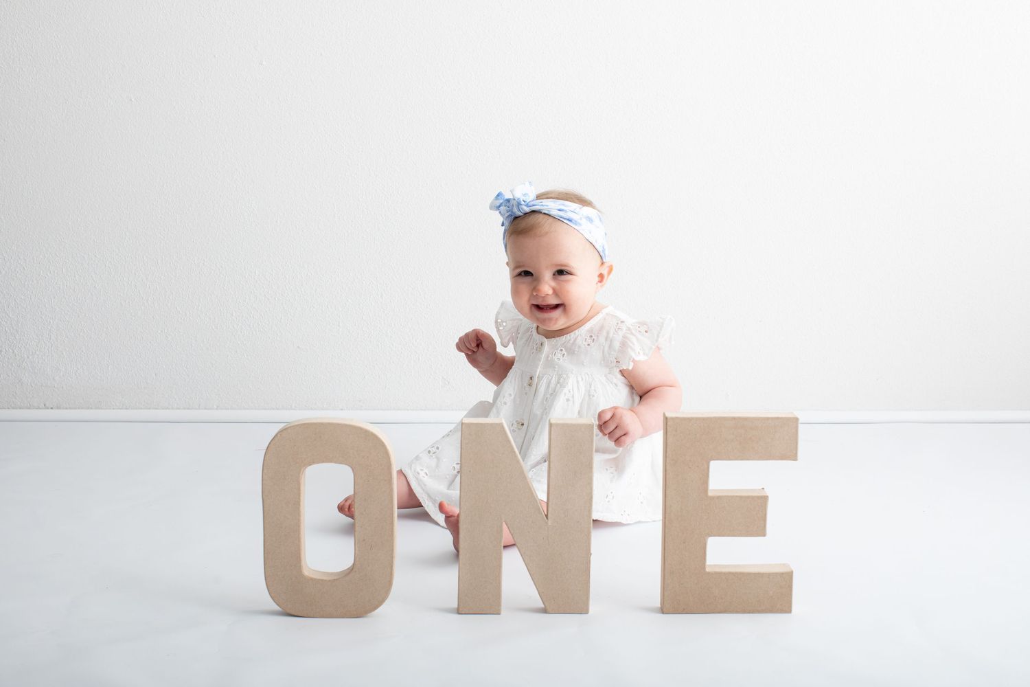Baby wearing blue headband sits beside large wooden letters spelling ONE on white background.