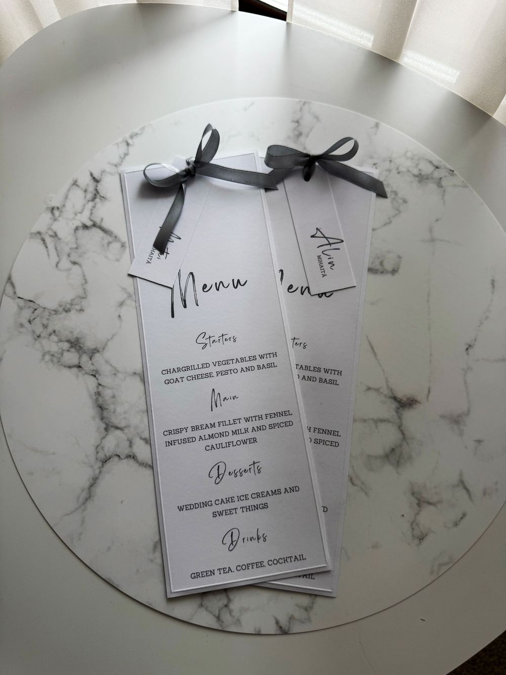 Elegant wedding menu cards with gray ribbon bows displayed on a marble table, featuring starters, main, desserts, and drinks.