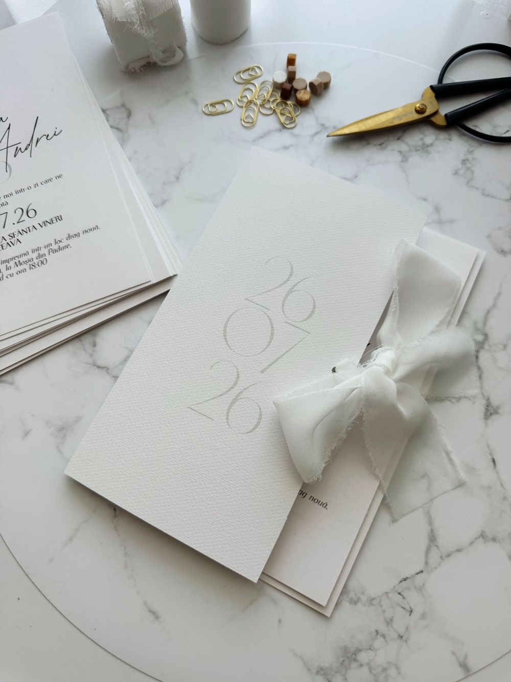 Elegant white embossed wedding invitation with silk bow on marble surface, surrounded by gold scissors and stationery.