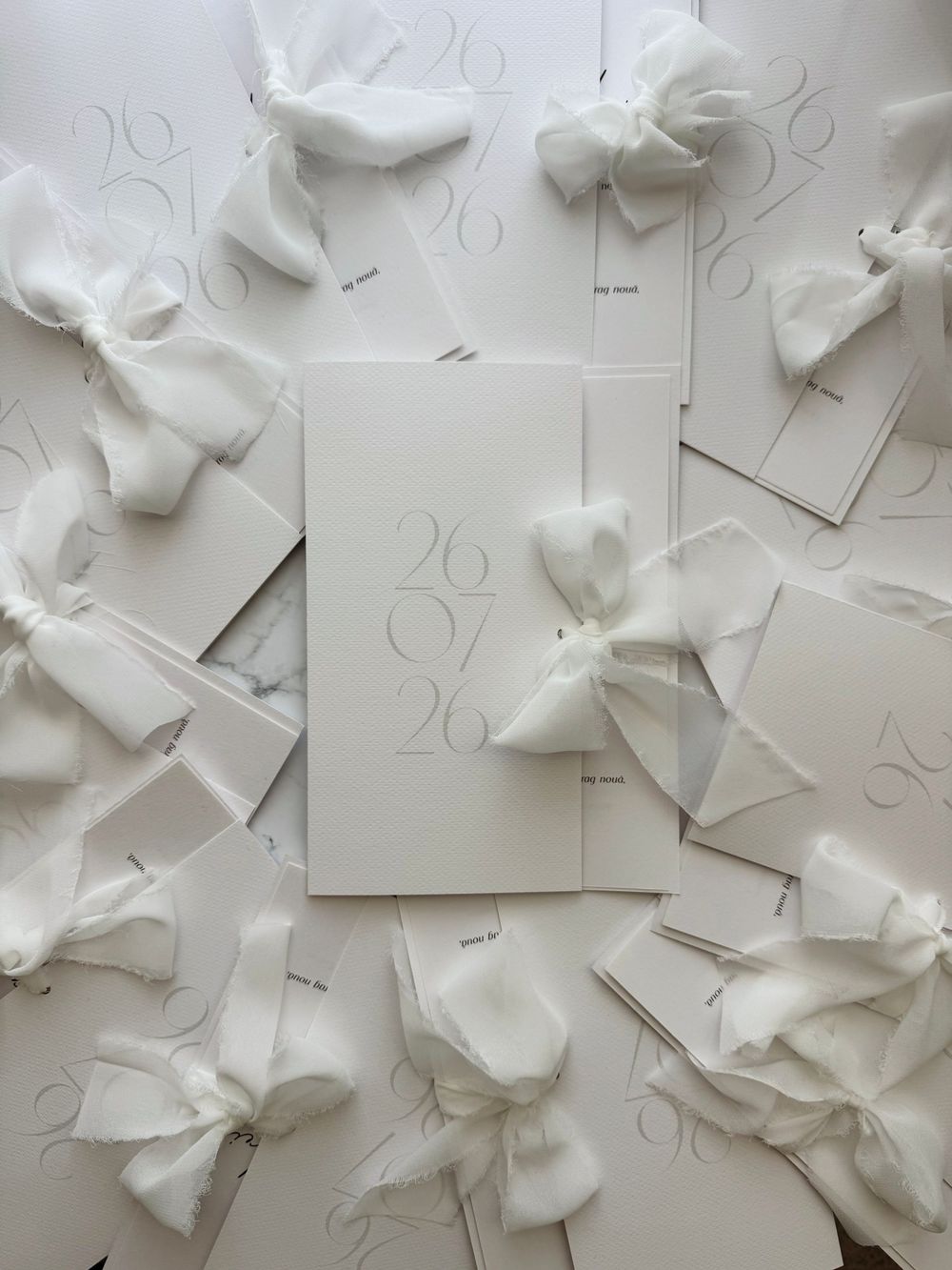 Elegant white wedding invitations with sheer ribbon bows and date 26/04/26 arranged on marble surface.