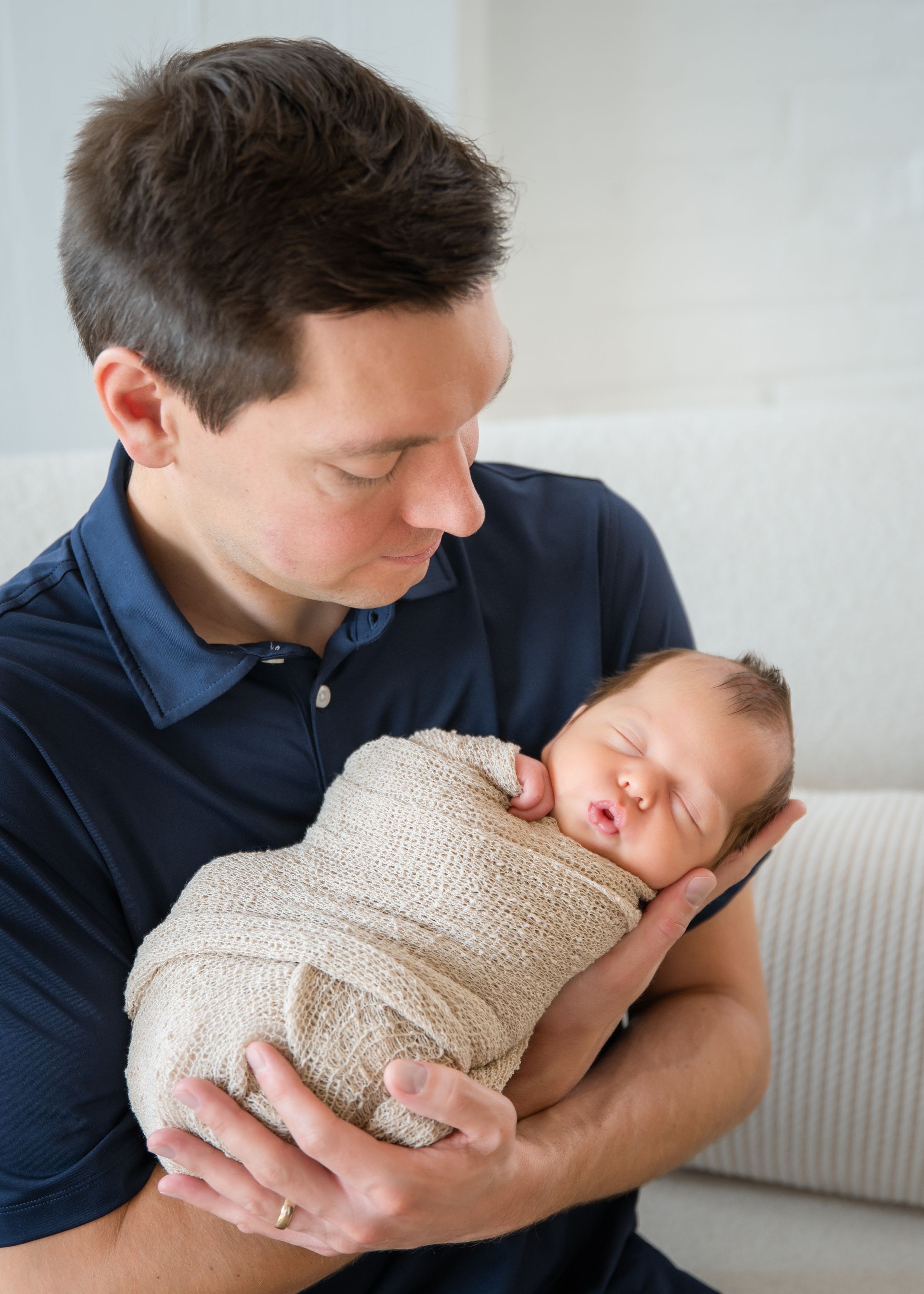 A person in a navy shirt tenderly holds and kisses a sleeping newborn baby wrapped in a knit blanket.